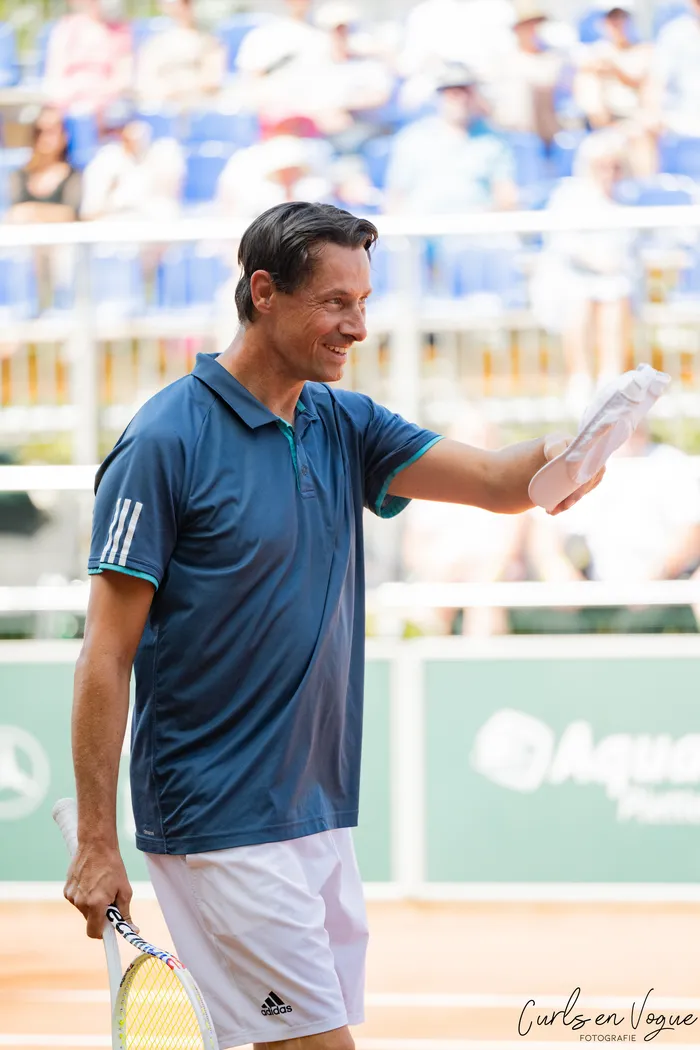 Ein lächelnder Mann in Tennisbekleidung hält einen Schläger und ein weißes Tuch auf einem Sandplatz bei den Platzmann Open 2026.