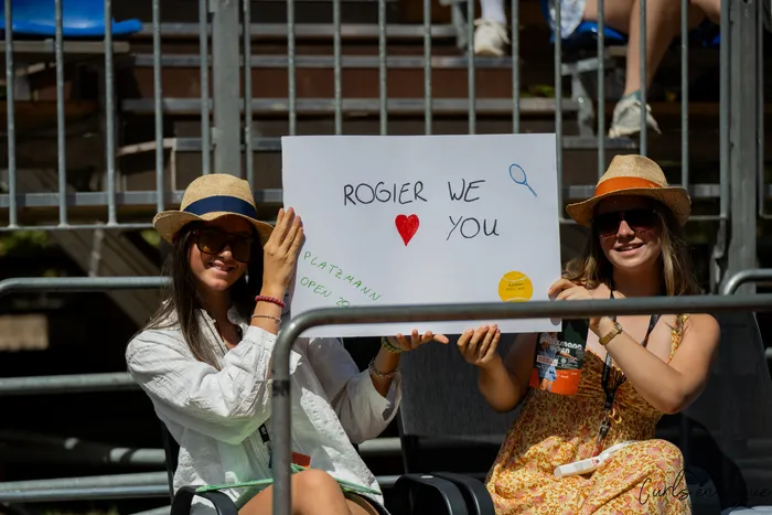 Zwei Frauen mit Strohhüten und Sonnenbrillen halten ein Schild mit der Aufschrift „ROGIER WE ❤️ YOU“ und „PLATZMANN OPEN 2026“.