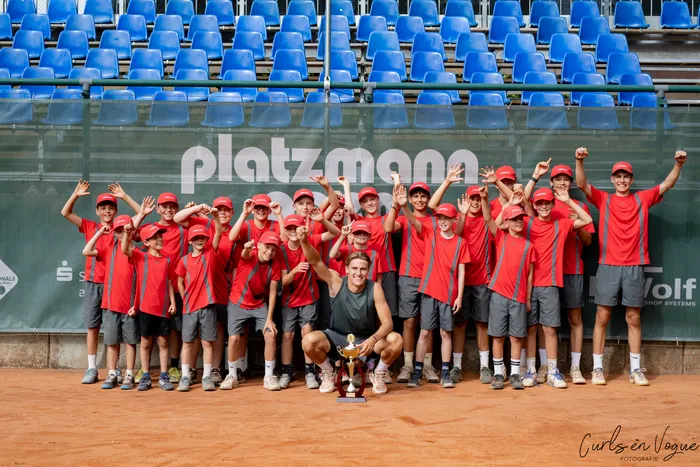 Eine Gruppe junger Männer in roten Trikots feiert auf einem Sandplatz vor blauen Tribünen, während ein Mann in der Mitte eine Trophäe hält.