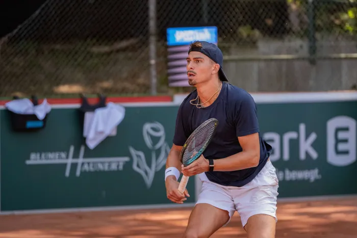 Ein junger Mann mit Baseballkappe und dunklem T-Shirt spielt Tennis auf einem roten Sandplatz.