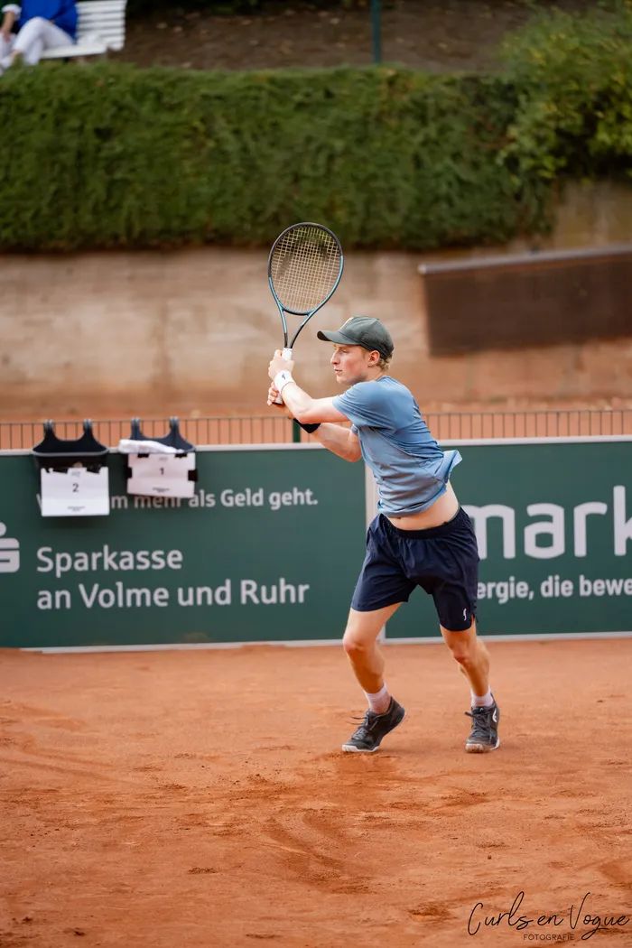 Ein Tennisspieler in blauem T-Shirt, dunklen Shorts und grüner Kappe bereitet sich auf einem Sandplatz auf einen Schlag vor.
