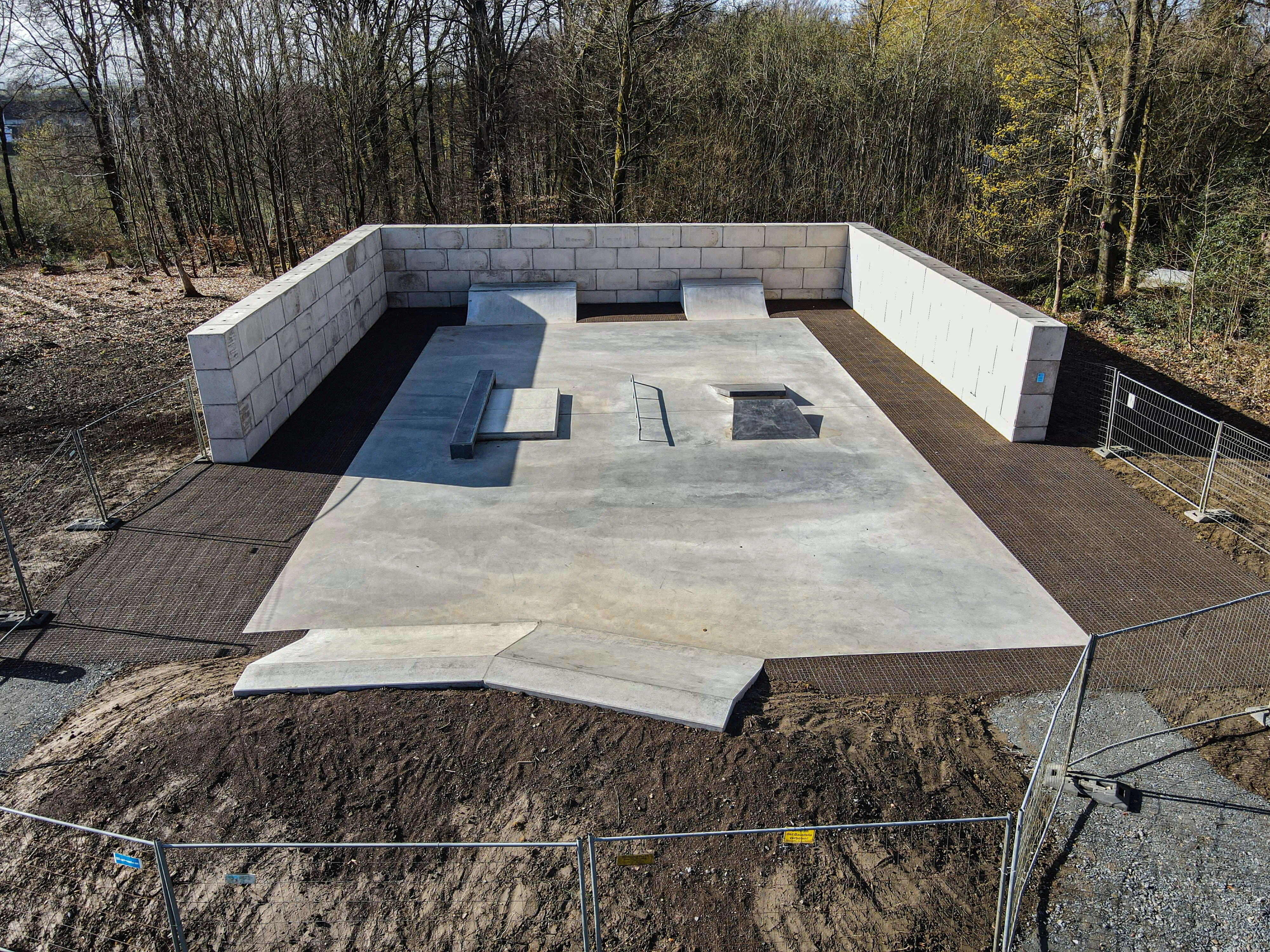Beton-Skatepark mit Rampen und Hindernissen, umgeben von Bauzäunen und freier Fläche.