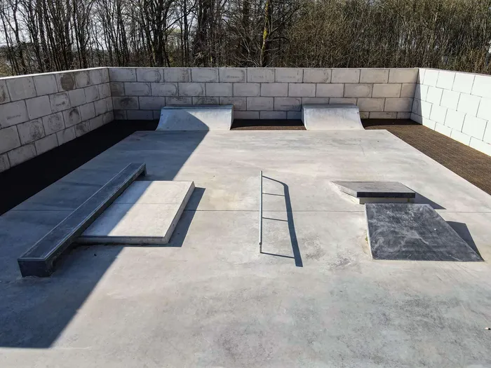 Betonierte Skateranlage mit verschiedenen Rampen, Rails und einer Halfpipe, umgeben von grauen Betonwänden. Im Hintergrund sind Bäume ohne Blätter zu sehen.