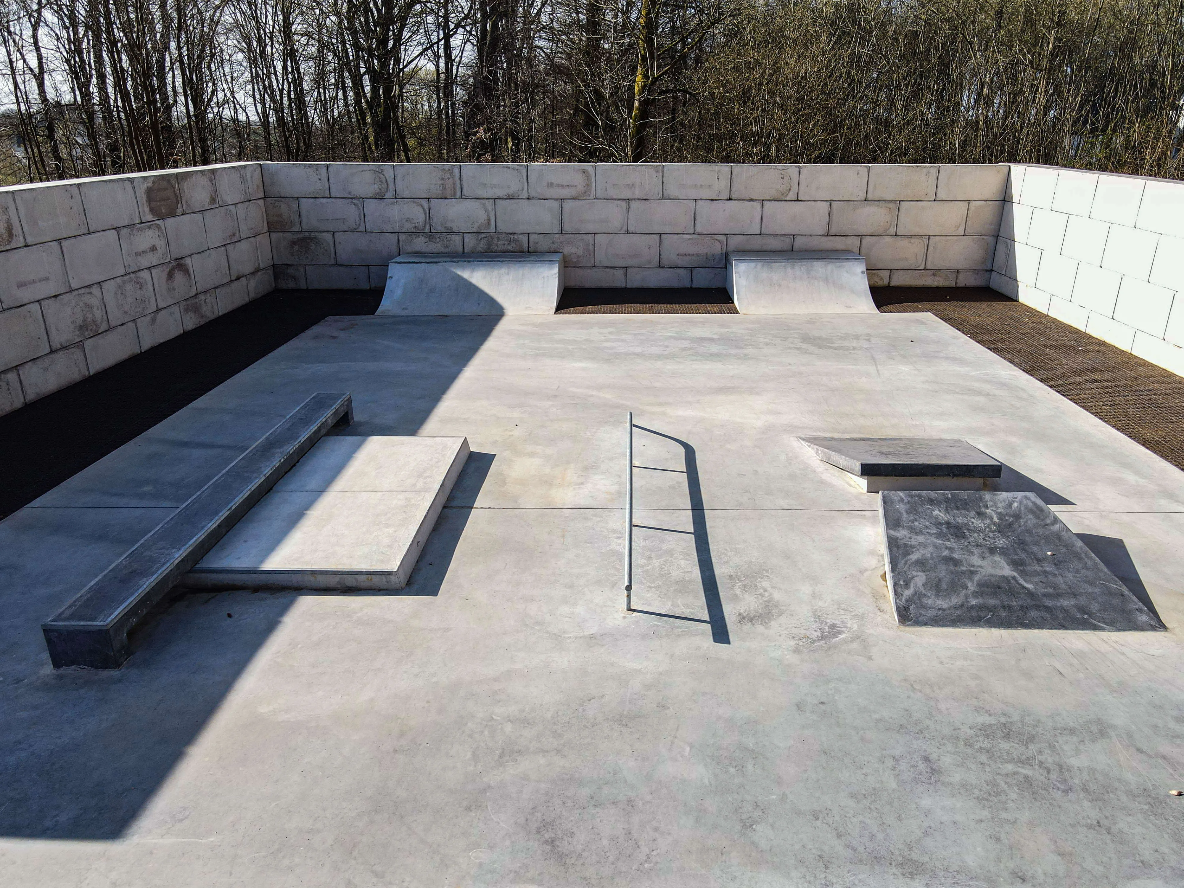 Beton-Skatepark mit verschiedenen Rampen und Hindernissen, umgeben von einer grauen Mauer und Bäumen im Hintergrund.