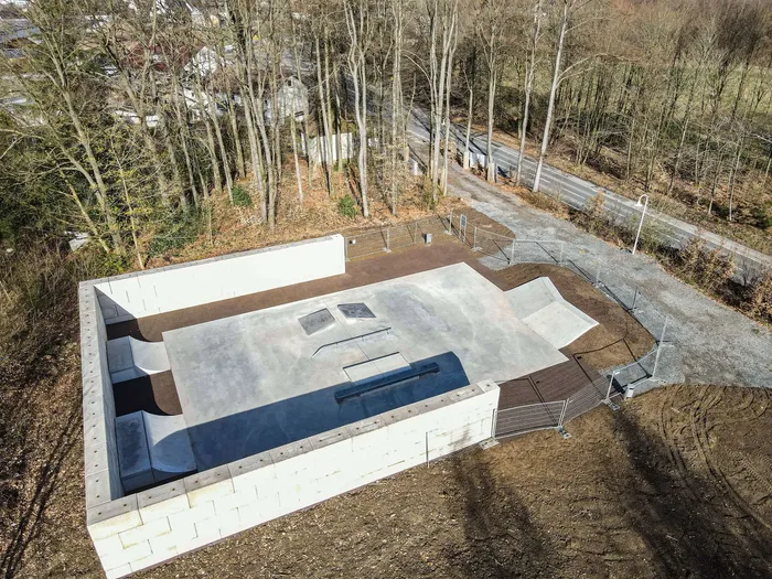 Ein Vogelperspektivenbild zeigt eine neu erbaute, rechteckige Skateranlage mit Rampen und einer Bank inmitten von Bäumen und angrenzend an eine Straße. Die Anlage ist von Betonmauern umgeben und wird durch ein Metallgeländer gesichert.