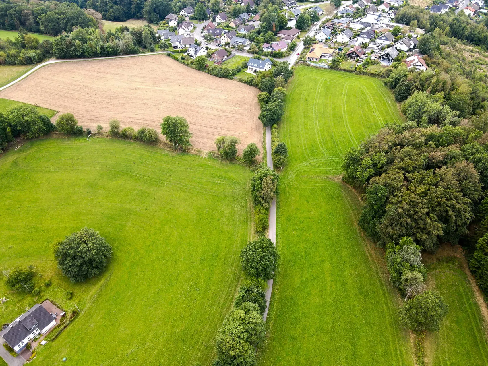 Luftaufnahme einer Landschaft mit grünen Wiesen, einem Acker und einem angrenzenden Wohngebiet.