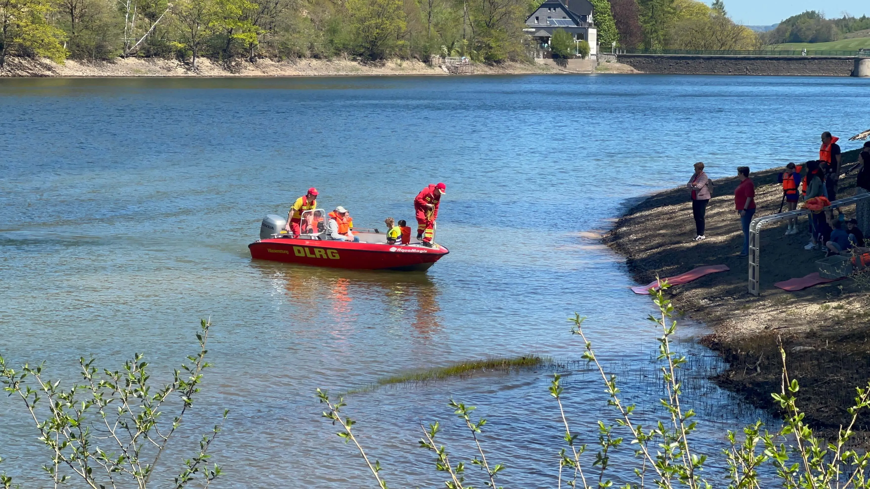Rotes DLRG-Boot mit mehreren Personen im Wasser vor Ufer mit Zuschauern an einem sonnigen Tag.