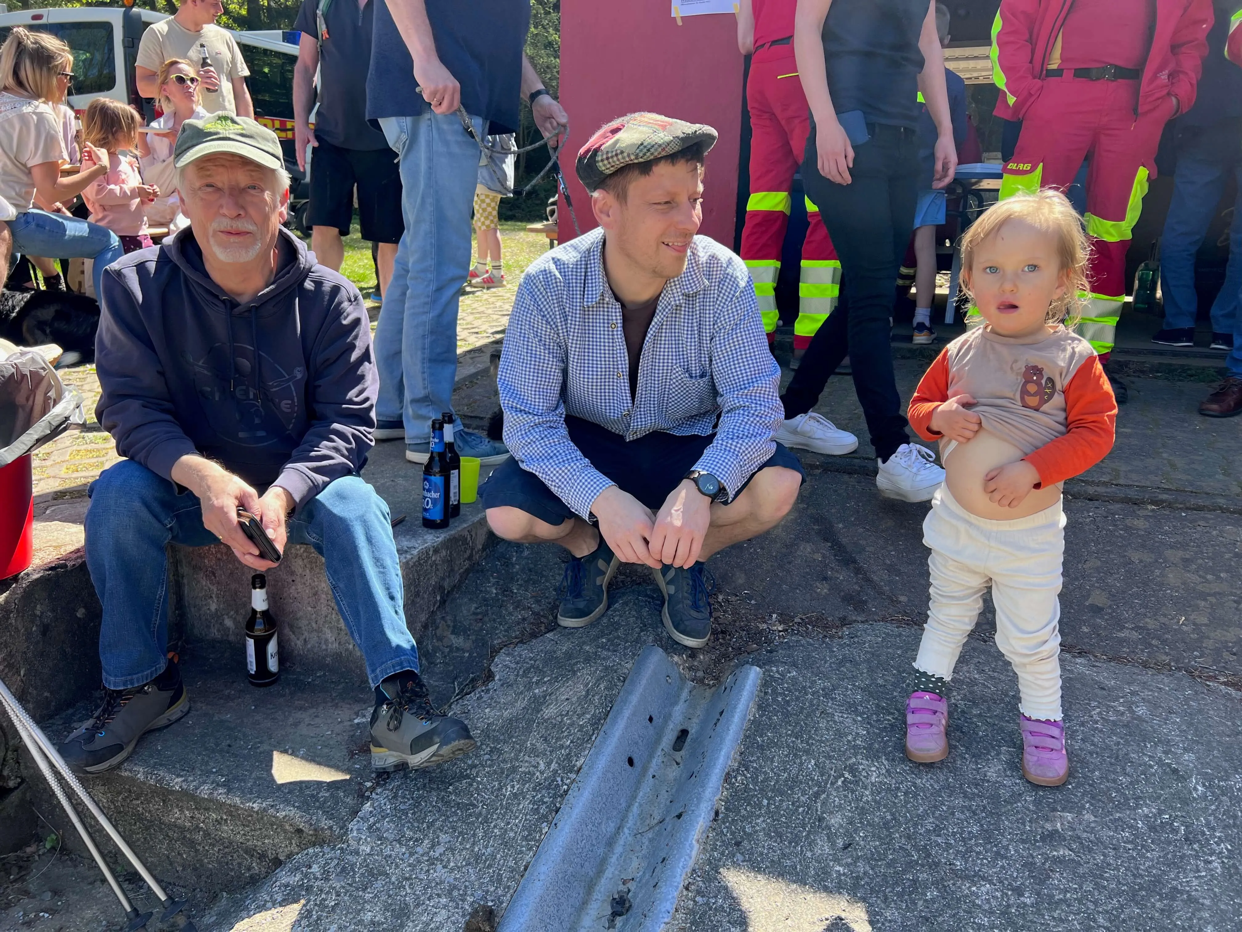 Zwei Männer und ein Kleinkind sitzen oder stehen an einem sonnigen Tag an einem Wasserrand. Im Hintergrund sind weitere Personen und ein rotes Einsatzfahrzeug zu sehen.