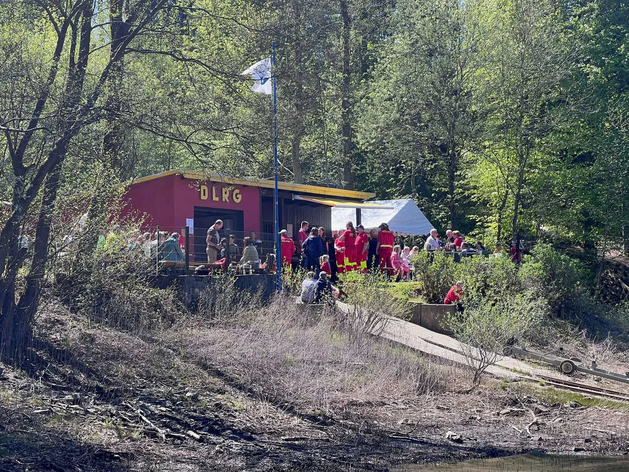 DLRG-Gebäude mit Zuschauern und Rettungsschwimmern in roten Westen an der Oestertalsperre, im Hintergrund Bäume und ein Zelt.