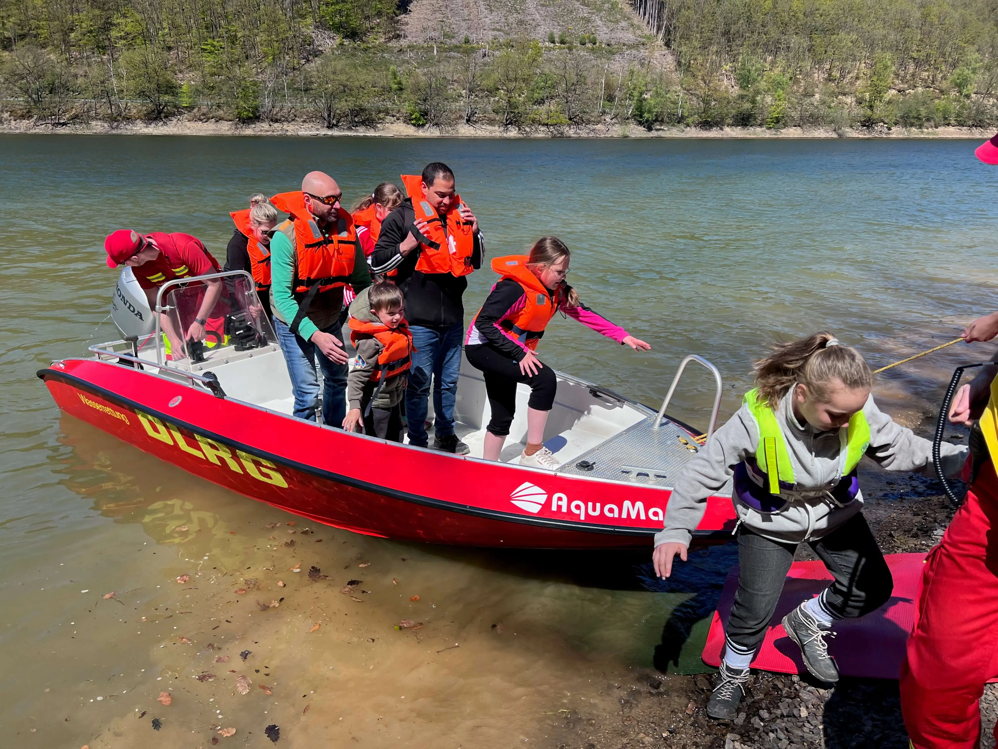 Rotes Rettungsboot mit mehreren Personen in Schwimmwesten wird an Land gezogen, während ein Mädchen vom Ufer aus hilft.
