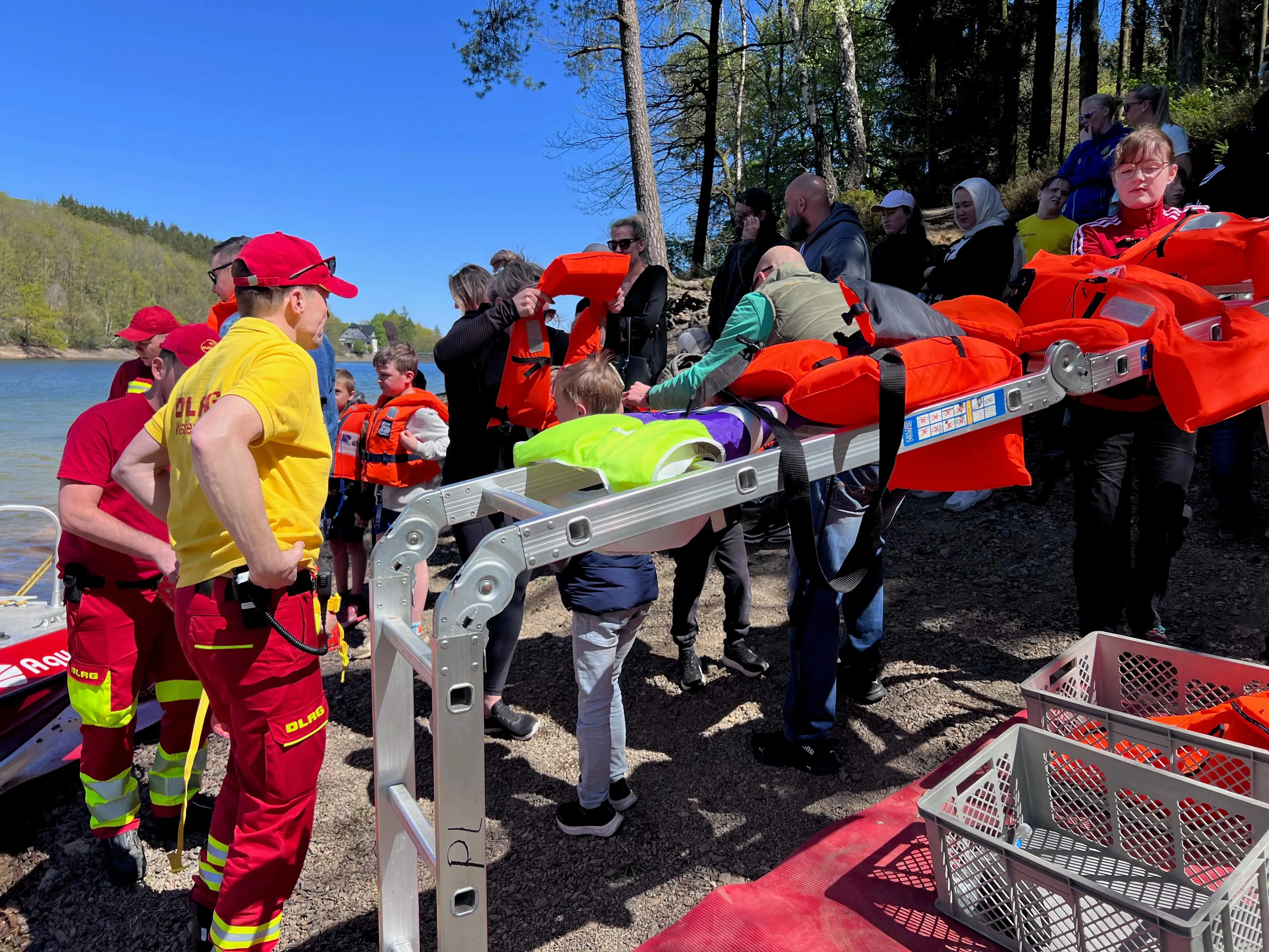 Rettungskräfte der DLRG tragen eine Person auf einer Trage vom Ufer in ein Boot. Umstehende beobachten die Aktion, viele tragen Schwimmwesten.