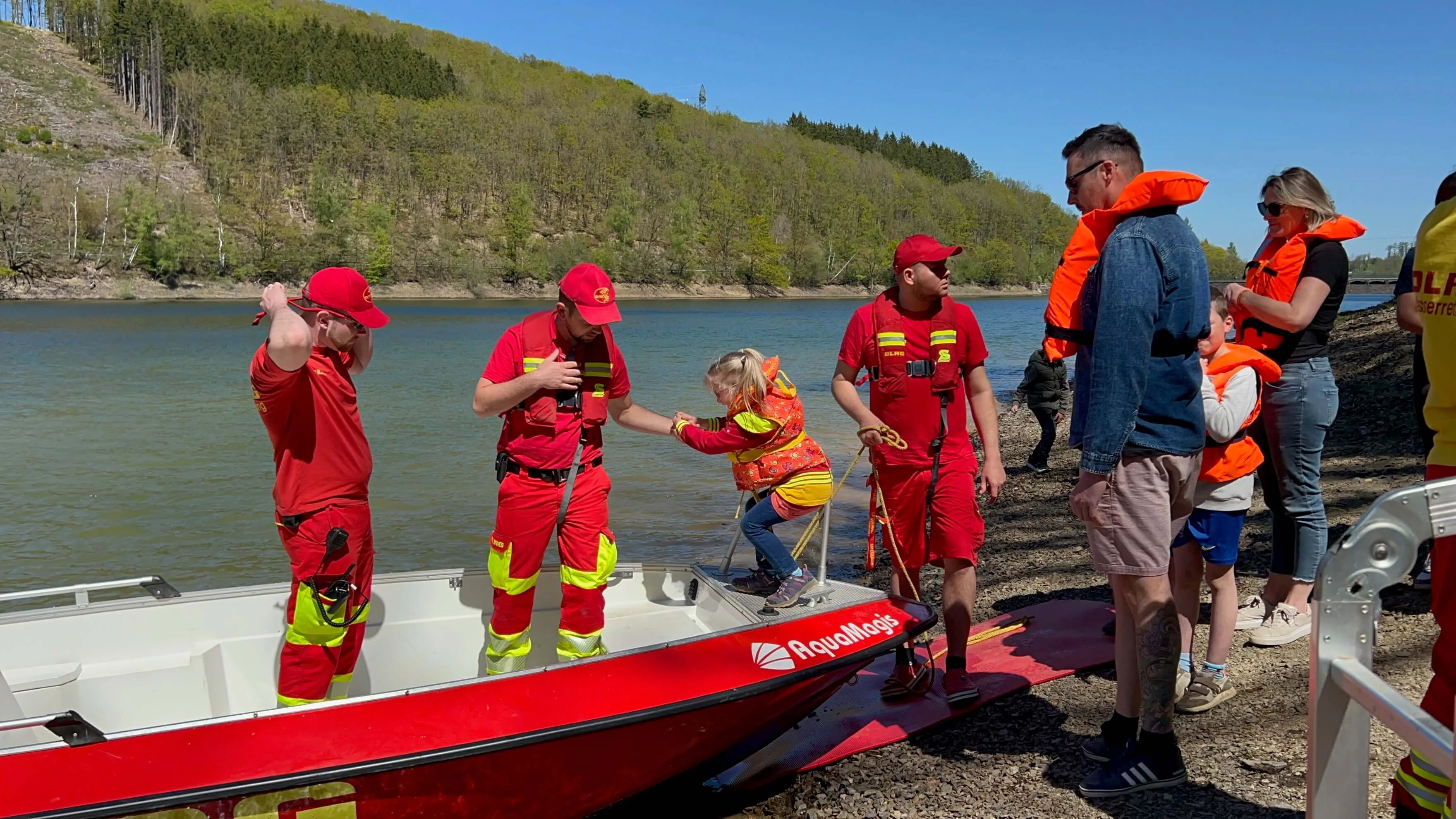 Feuerwehrleute helfen einem Kind beim Anlegen einer Schwimmweste am Ufer der Oestertalsperre, während andere Besucher zusehen.
