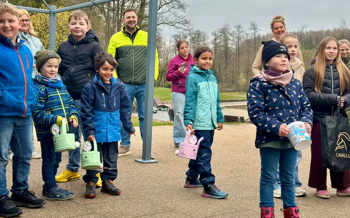 Eine Gruppe Kinder hält Ostereierkörbe bereit, während sie auf einem Gelände mit Bäumen und einem kleinen See stehen. Im Hintergrund sind weitere Personen und ein metallener Bogen zu sehen.
