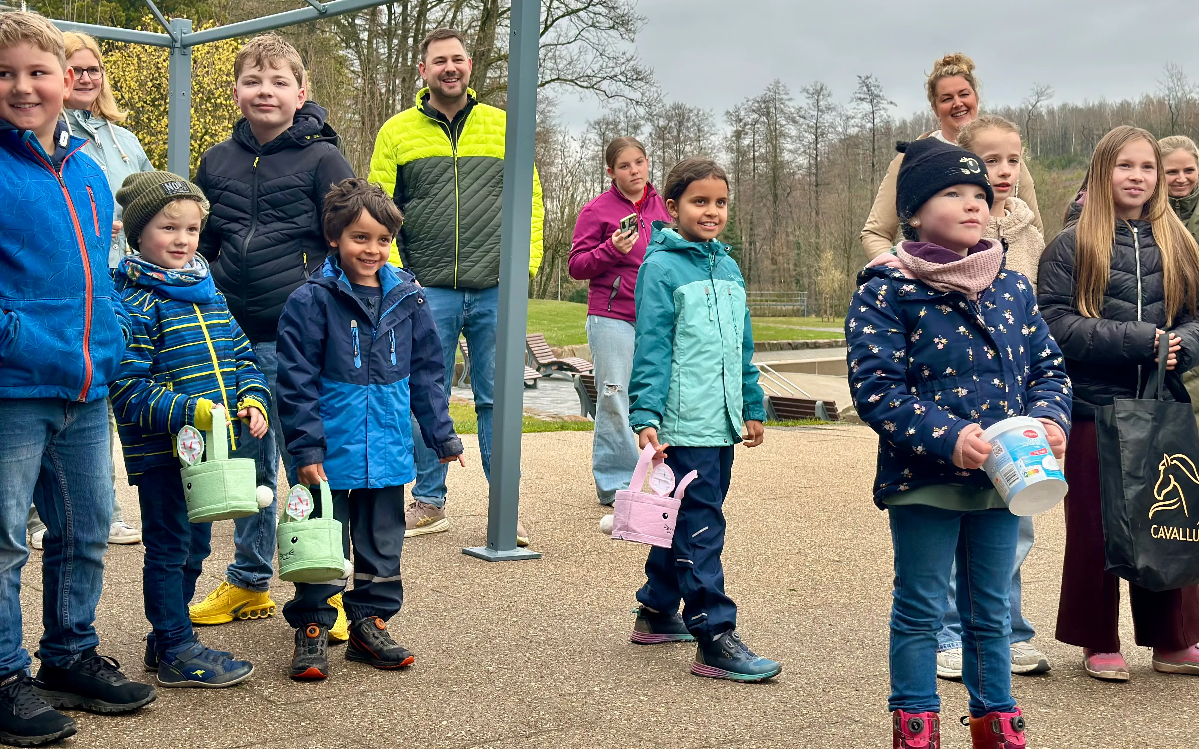 Eine Gruppe Kinder hält Ostereierkörbe bereit, während sie auf einem Gelände mit Bäumen und einem kleinen See stehen. Im Hintergrund sind weitere Personen und ein metallener Bogen zu sehen.