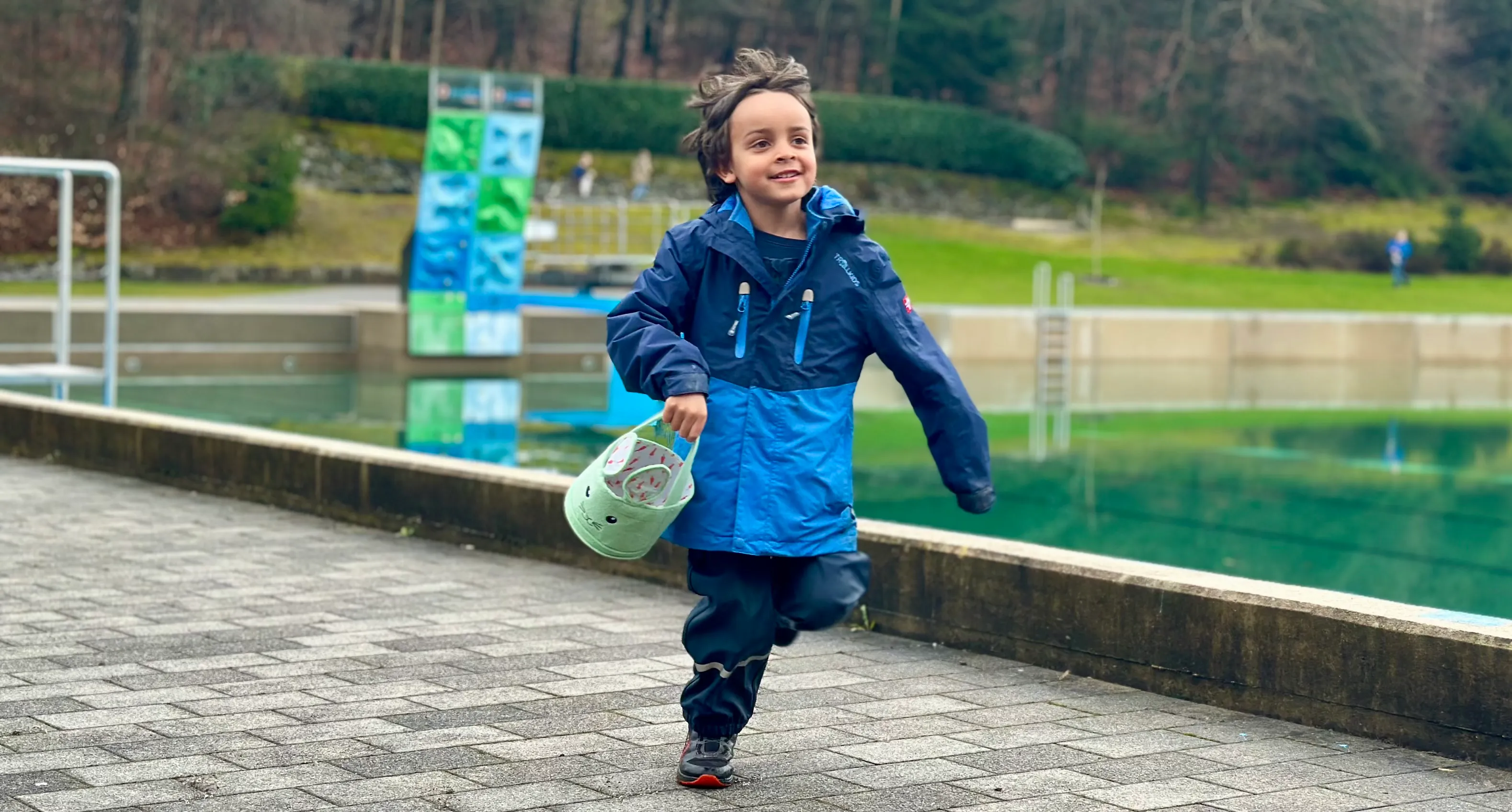Ein Kind rennt lächelnd mit einem Eimer auf einem gepflasterten Weg, im Hintergrund ein Fluss und Bäume.