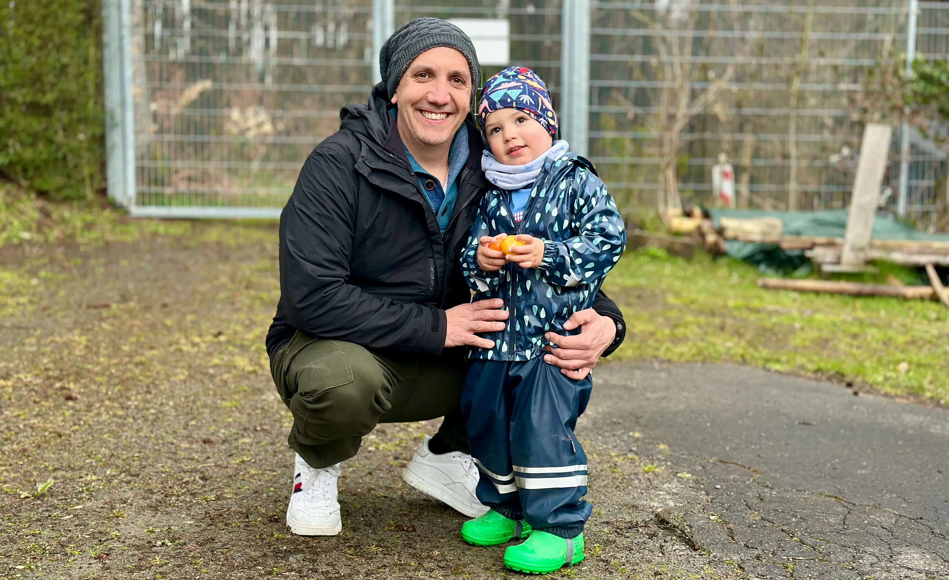 Ein lächelnder Mann kauert neben einem kleinen Kind mit Mütze und Regenkleidung, das ein Osterei hält. Im Hintergrund ist ein Zaun und Vegetation zu sehen.