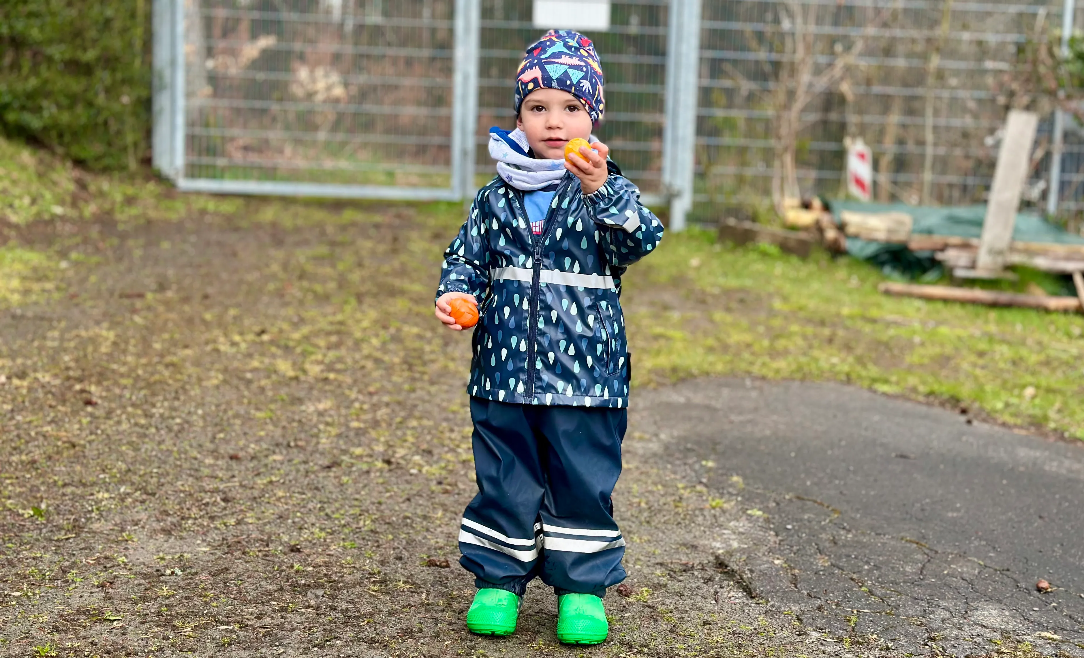 Ein Kind in wetterfester Kleidung hält zwei Mandarinen in den Händen, während es auf einem Kiesweg vor einem Zaun steht.