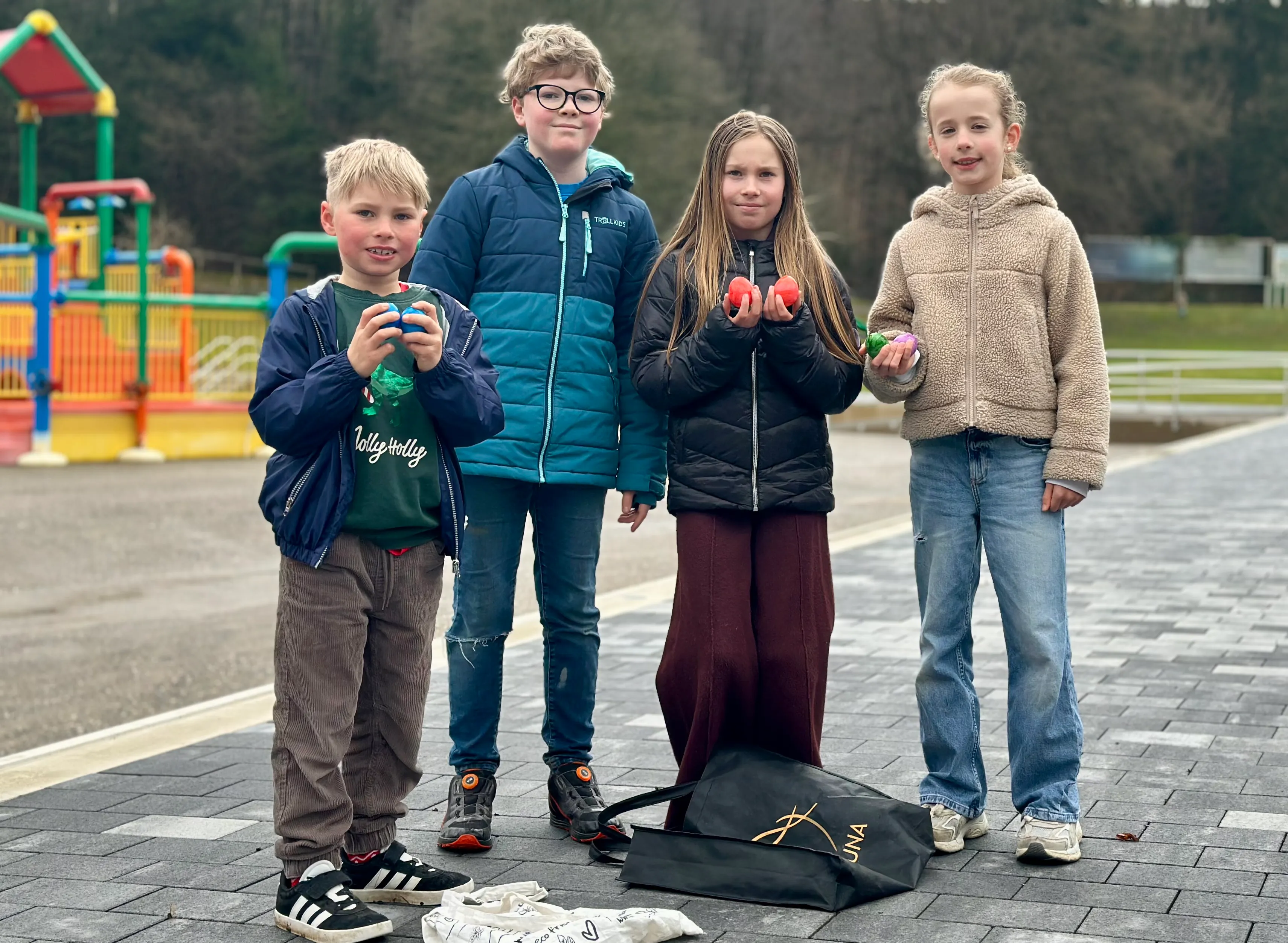 Vier Kinder stehen auf einem gepflasterten Weg vor einem Spielplatz und halten Ostereier in den Händen. Eine Tasche mit dem DLRG-Logo steht vor ihnen.