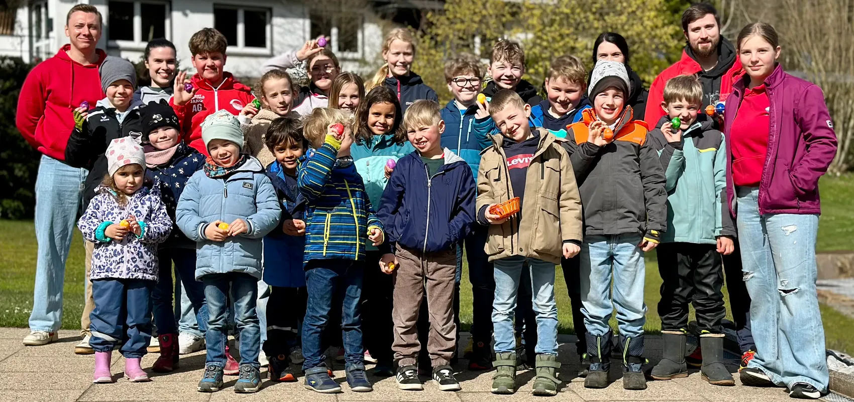 Eine Gruppe von Kindern und Betreuern der DLRG Halver steht lächelnd mit Ostereiern in den Händen vor einem Gebäude und einem grünen Hintergrund zusammen.