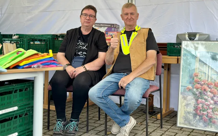Zwei Personen sitzen unter einem Zelt; der Mann gibt ein Daumen-hoch-Zeichen und trägt eine gelbe Warnweste um den Hals.