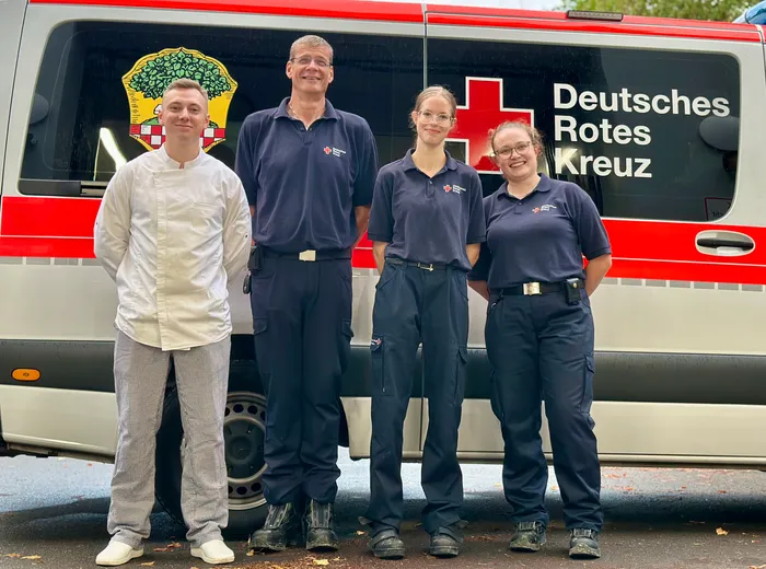Ein Koch in weißer Arbeitskleidung und drei Einsatzkräfte des Deutschen Roten Kreuzes in dunkelblauen Uniformen stehen vor einem DRK-Fahrzeug.