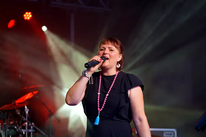 Eine Frau mit kurzen, roten Haaren singt in ein Mikrofon auf einer Bühne vor einem roten und weißen Lichtschein. Sie trägt ein schwarzes Oberteil, eine lange rosa Kette und große Ohrringe.