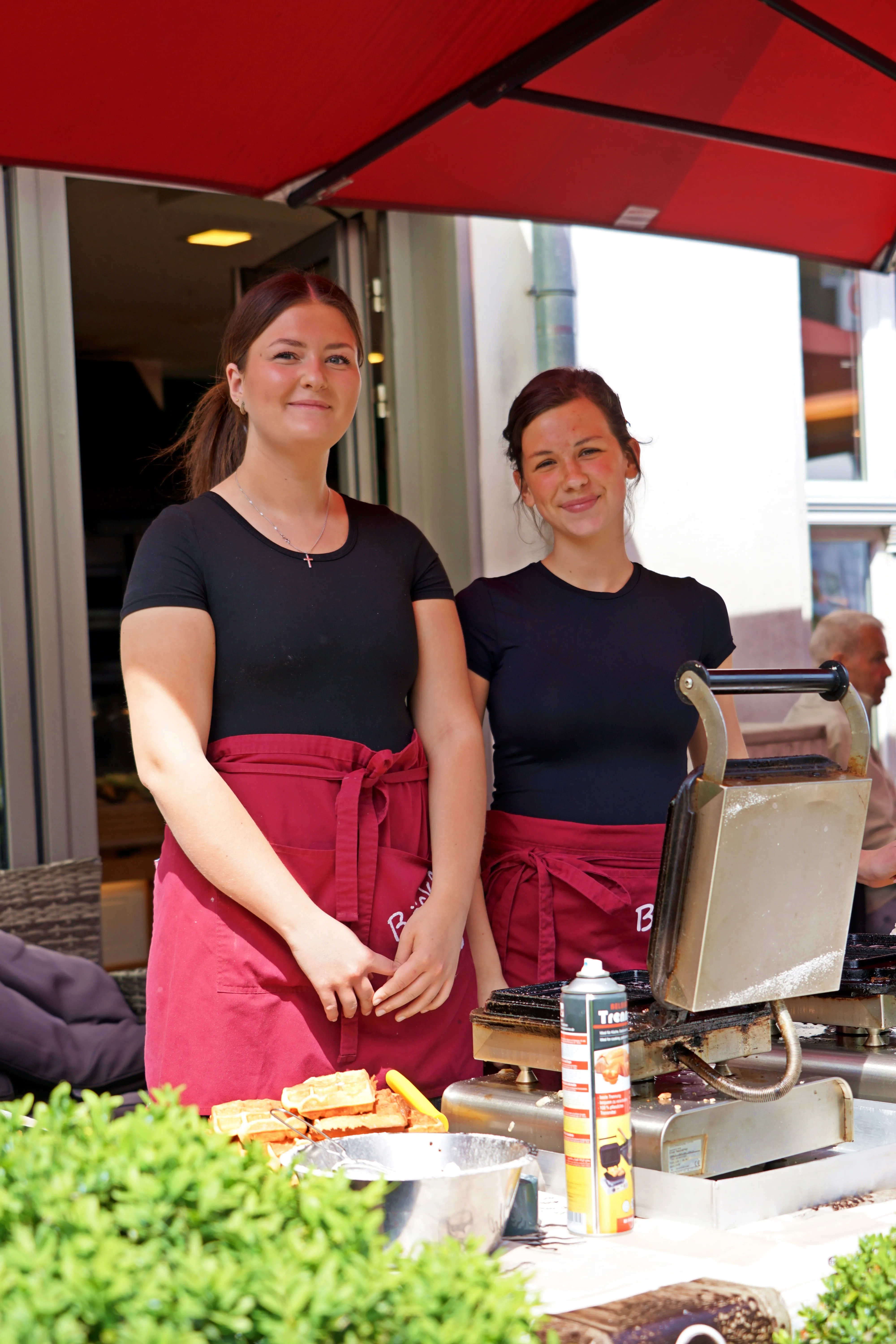 Zwei junge Frauen mit roten Schürzen stehen lächelnd neben einem Grillwagen und bereiten Speisen zu. Im Hintergrund ist ein belebter Platz zu sehen.