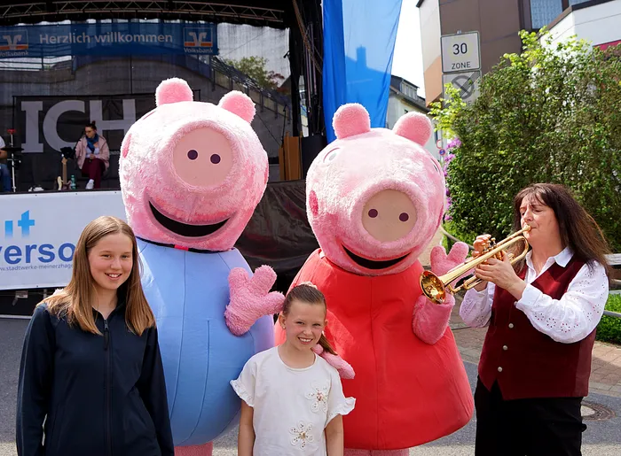 Zwei Personen in Peppa-Wutz-Kostümen stehen mit zwei Mädchen und einer Trompeterin vor einer Bühne mit dem Schriftzug „Meinerzhagen“.