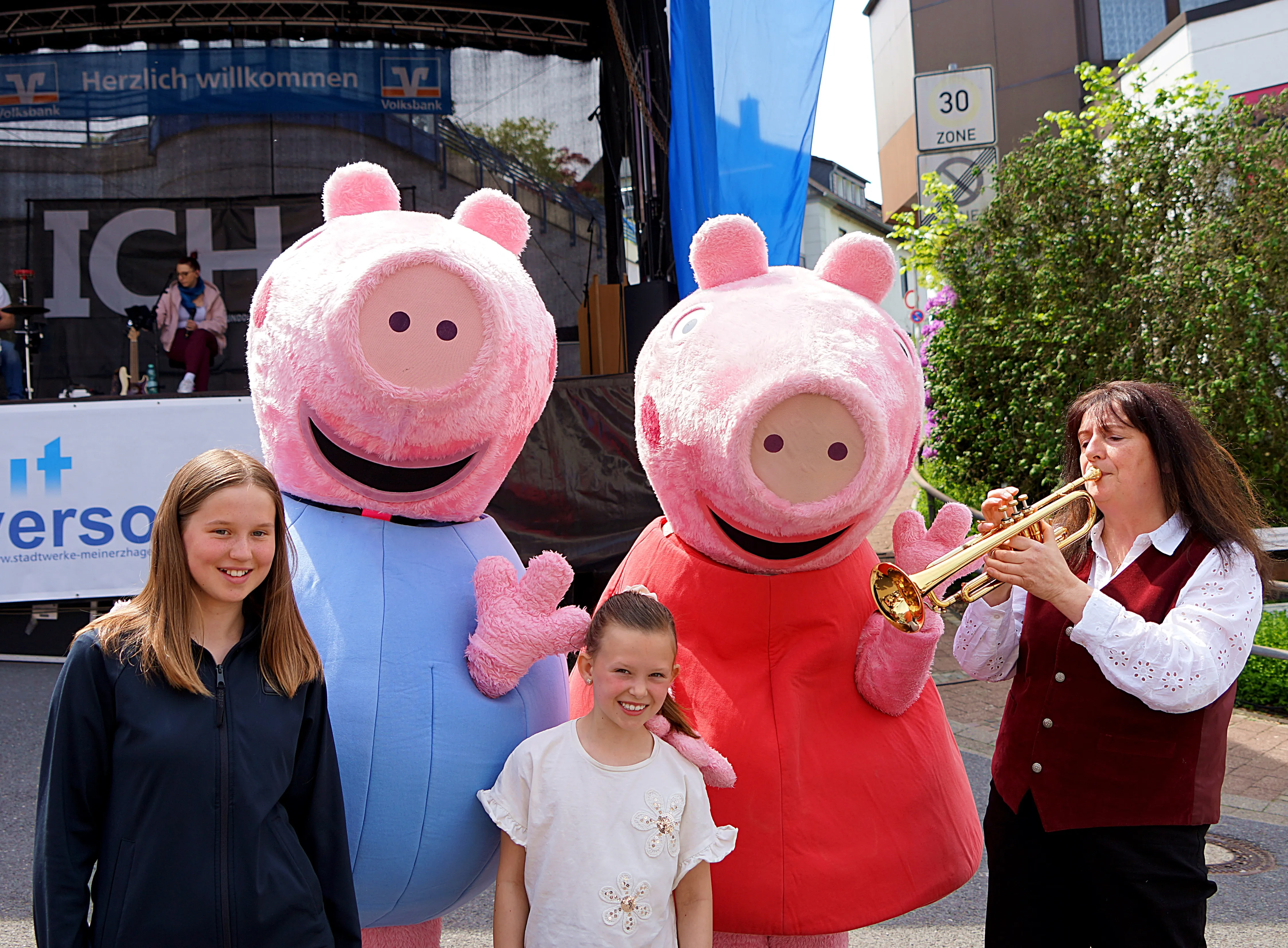 Zwei Personen in Peppa-Wutz-Kostümen stehen mit zwei Mädchen und einer Trompeterin vor einer Bühne mit dem Schriftzug „Meinerzhagen“.