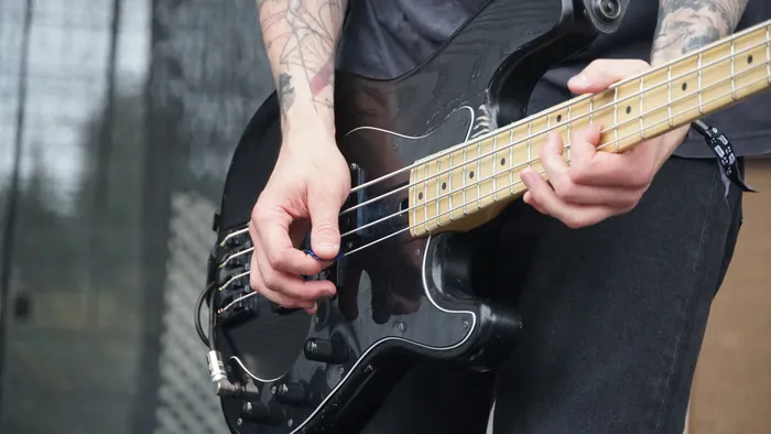 Eine tätowierte Person spielt einen schwarzen E-Bass mit hellem Griffbrett.
