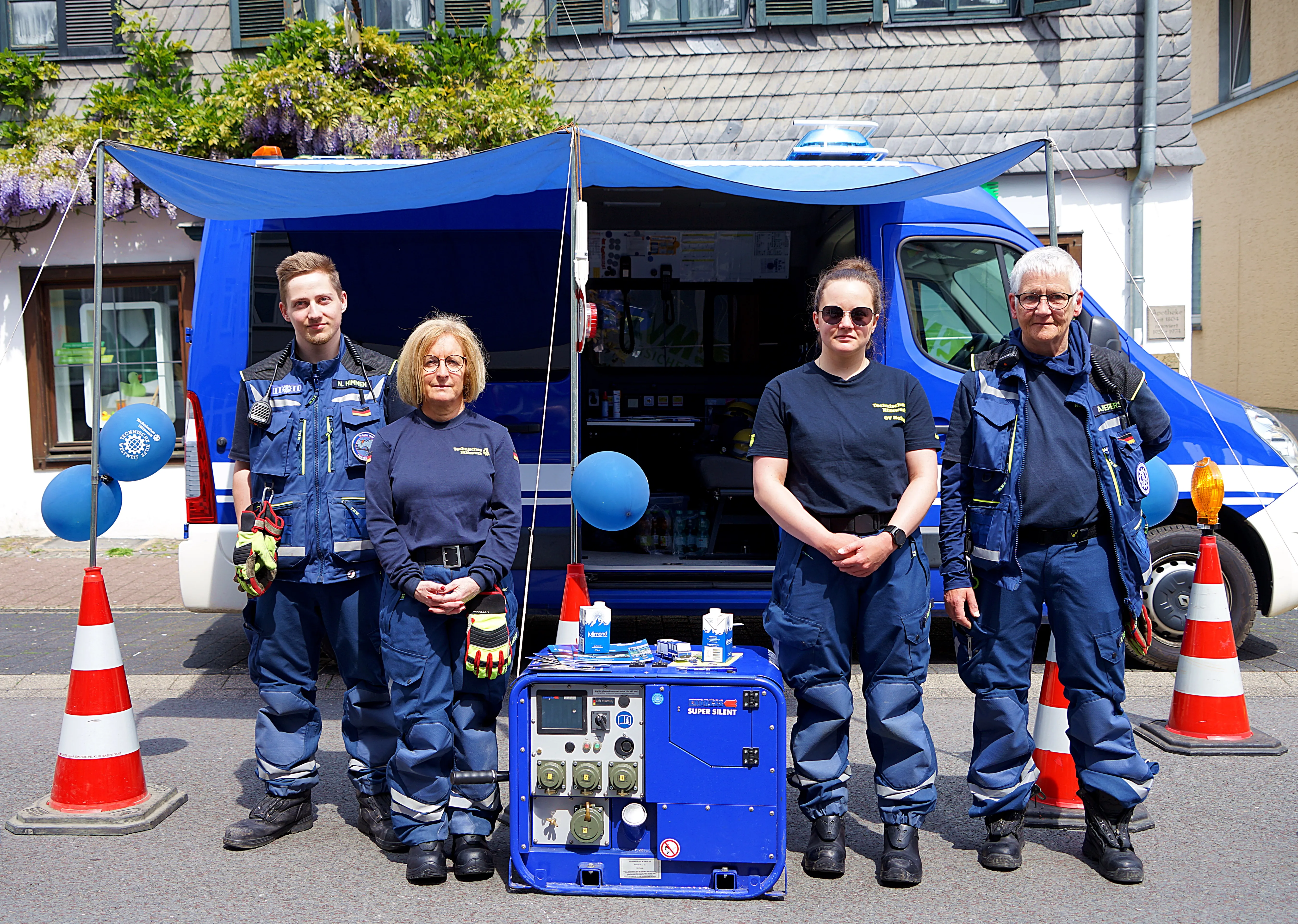 Vier Einsatzkräfte des Deutschen Roten Kreuzes stehen vor einem geöffneten Mannschaftswagen. Vor dem Wagen steht ein Generator und Dekoration in Form von blauen Luftballons und Verkehrskegeln.