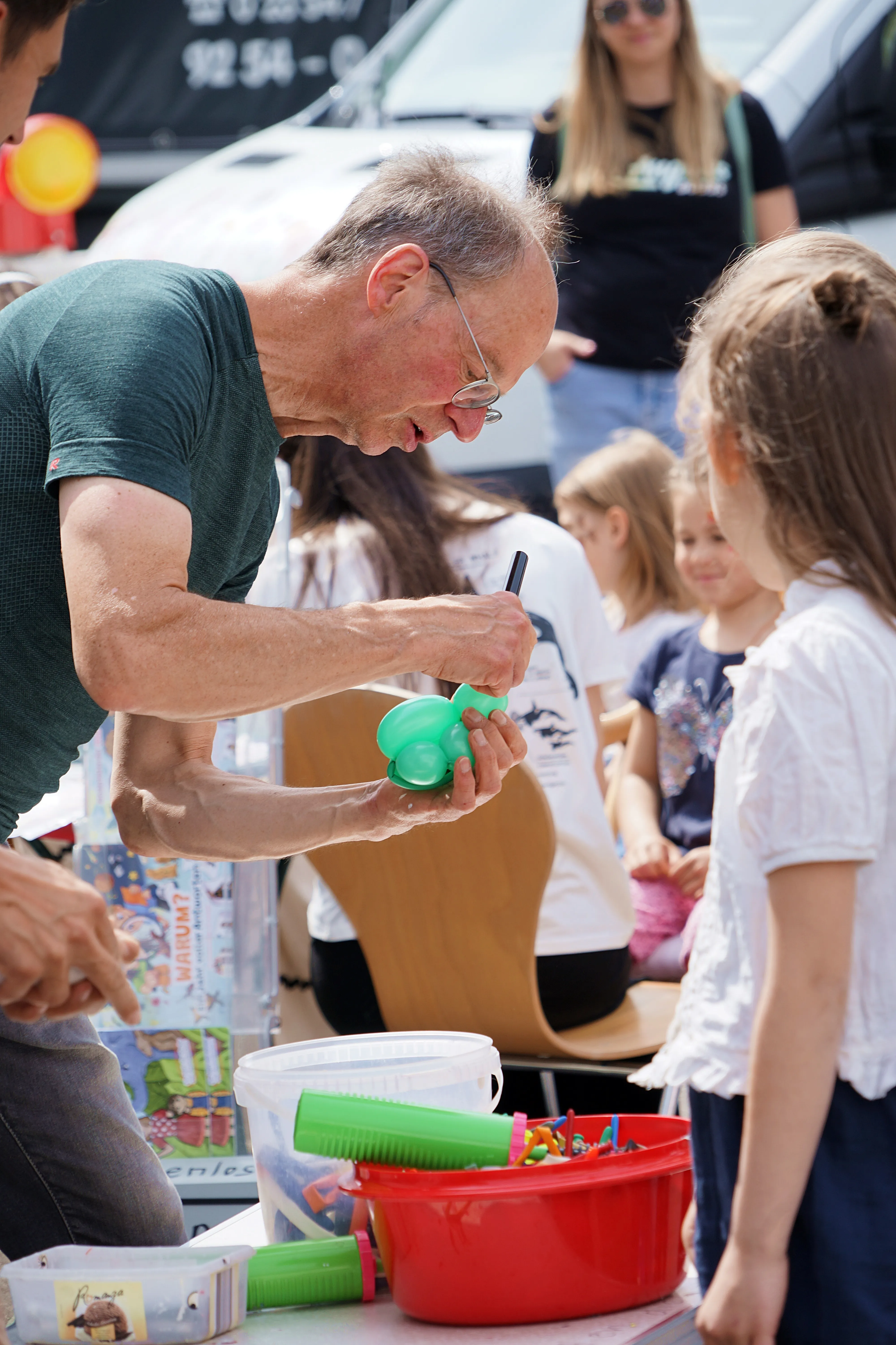 Ein älterer Mann mit Brille bastelt mit einem Mädchen an einem grünen Spielzeug. Im Hintergrund stehen weitere Menschen und ein Tisch mit Bastelmaterialien.