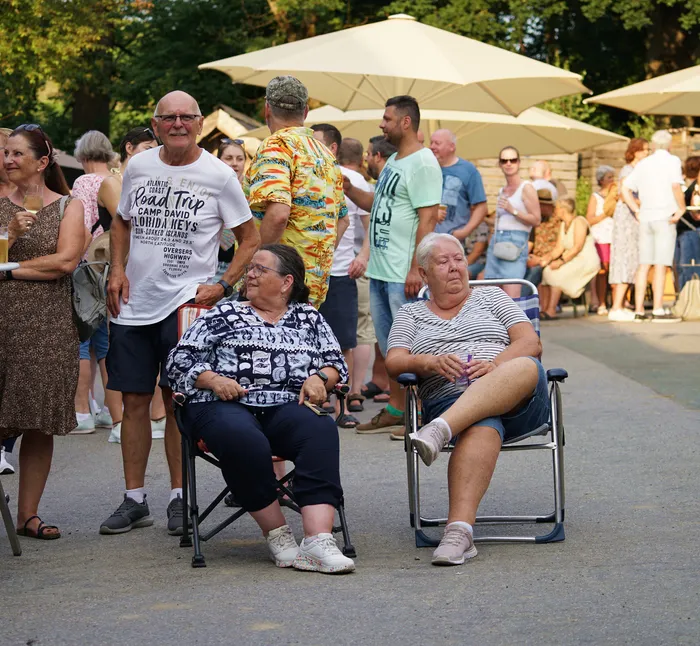 Zwei ältere Frauen sitzen auf Klappstühlen an einer Straße, umgeben von Zuschauern bei einer Veranstaltung im Freien. Im Hintergrund sind weitere Menschen und Sonnenschirme zu sehen.