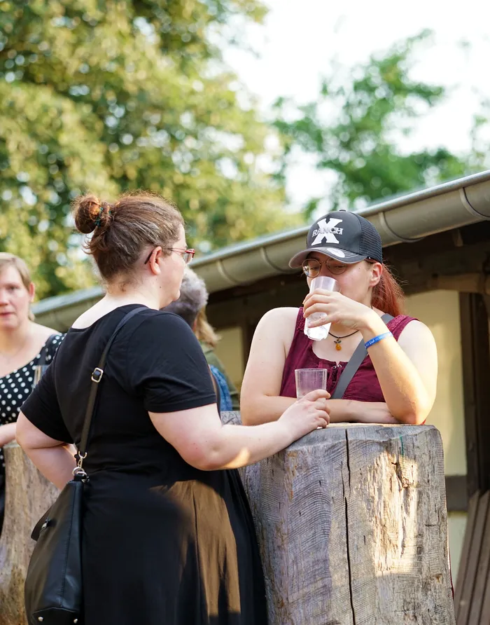 Zwei Frauen unterhalten sich im Freien, eine von ihnen lehnt an einem Baumstumpf und hält ein Getränk in der Hand. Im Hintergrund sind weitere Personen und ein Gebäude zu sehen.