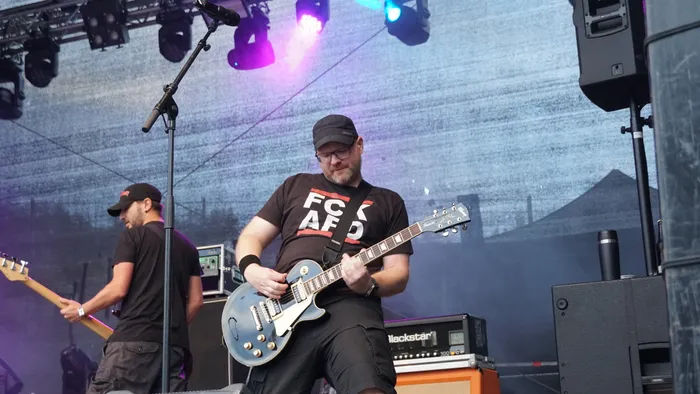 Ein Musiker mit Brille und schwarzer Kappe spielt eine schwarze E-Gitarre auf einer Bühne. Er trägt ein schwarzes T-Shirt mit der Aufschrift „FCK AFD“.