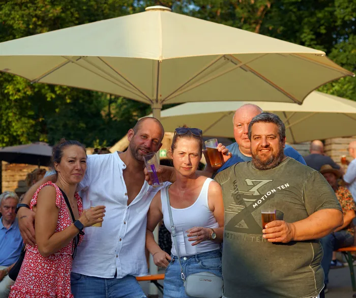 Fünf Menschen stehen lächelnd mit Getränken in der Hand vor Sonnenschirmen. Sie scheinen eine gesellige Zeit im Freien zu verbringen.