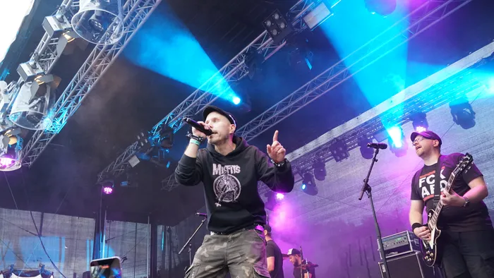 Ein Sänger in einem schwarzen Hoodie und Camouflage-Hosen singt unter blauem und violettem Bühnenlicht, während ein Gitarrist daneben spielt.