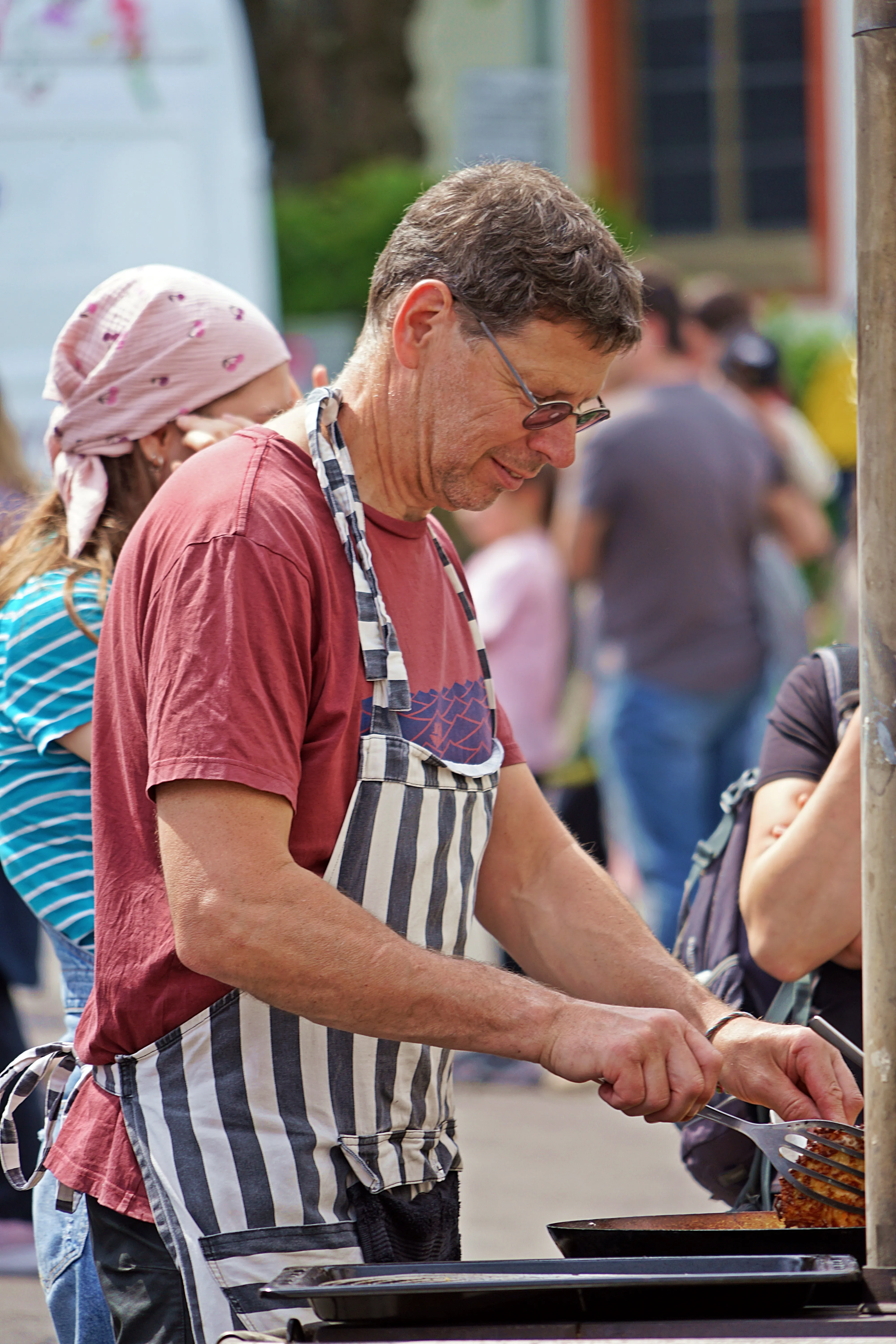 Mann mit Schürze brät auf einer Grillplatte Speisen auf einem Markt im Freien, während im Hintergrund Menschen vorbeigehen.