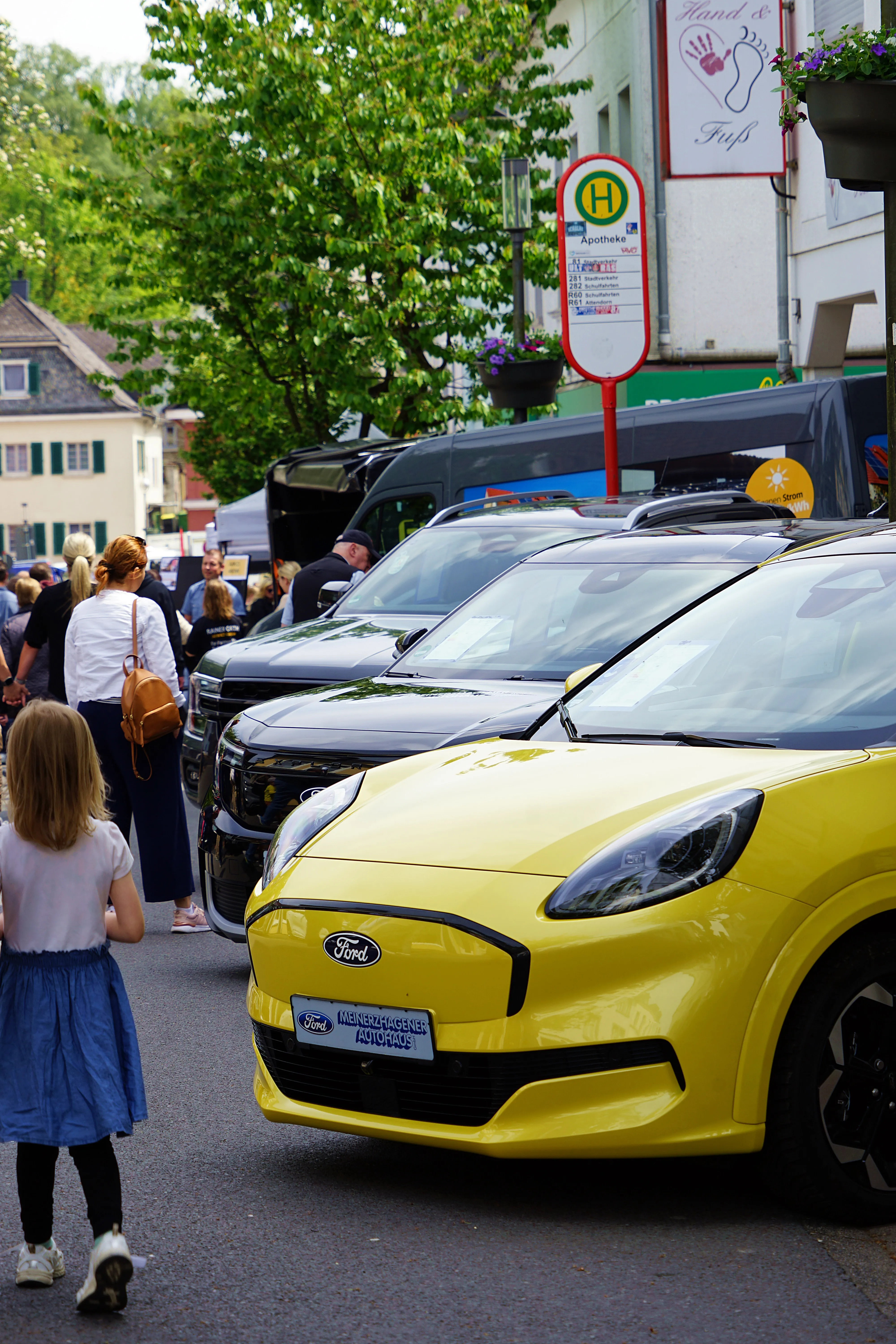 Gelber Ford Fiesta steht im Mittelpunkt einer Reihe von Autos auf einer belebten Straße, während Menschen im Hintergrund vorbeigehen. Ein Apotheken-Schild ist im Hintergrund zu sehen.