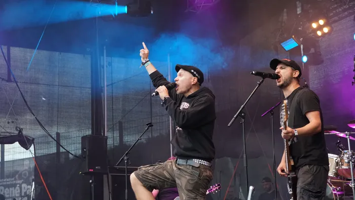 Ein Sänger in schwarzem Hoodie und Camouflage-Shorts singt mit erhobenem Finger, während ein Gitarrist daneben singt, unter blauem Bühnenlicht.