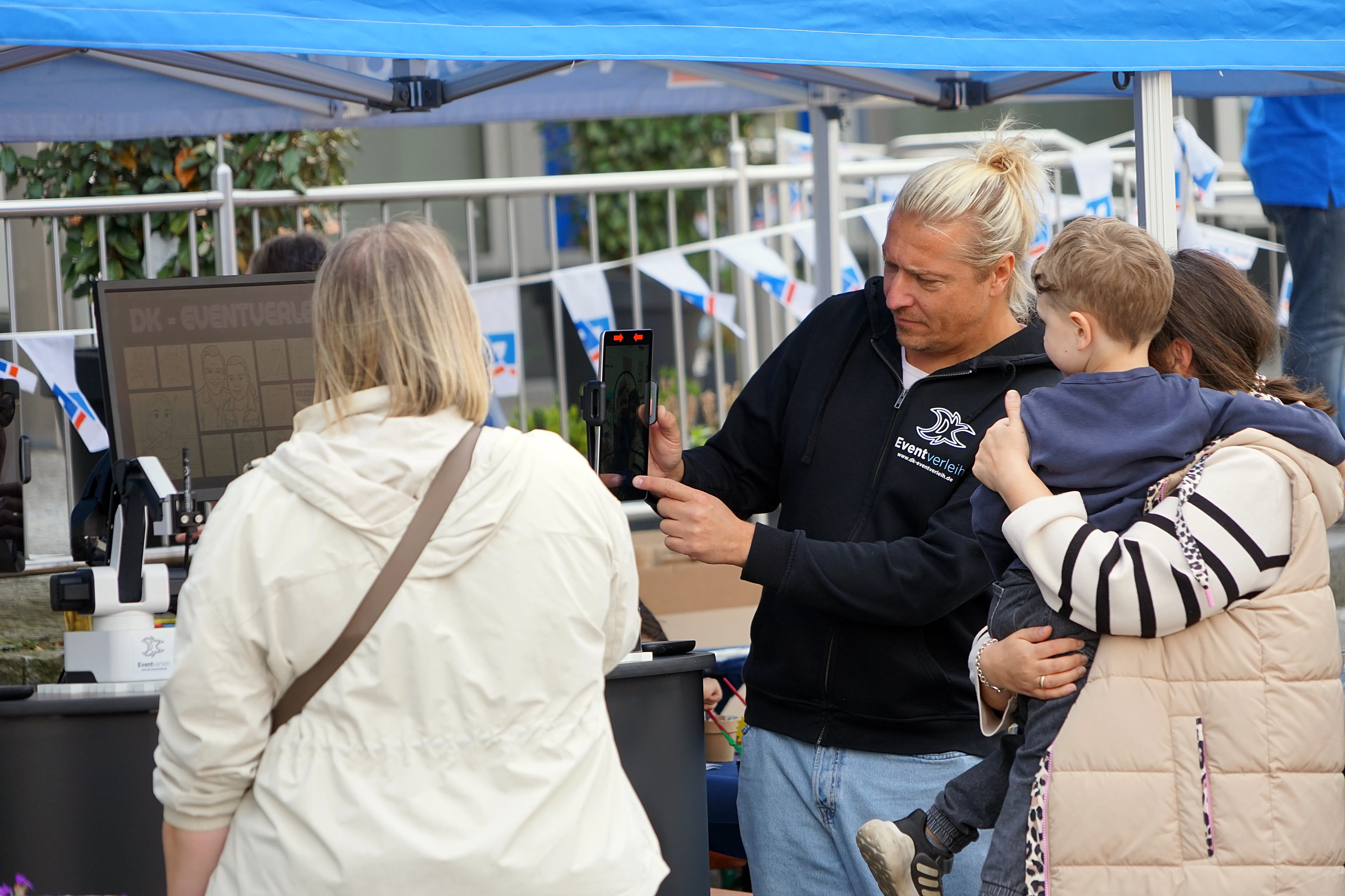 Ein Mann fotografiert mit seinem Smartphone eine Frau und ein Kind auf einem Stadtfest unter einem blauen Pavillon.