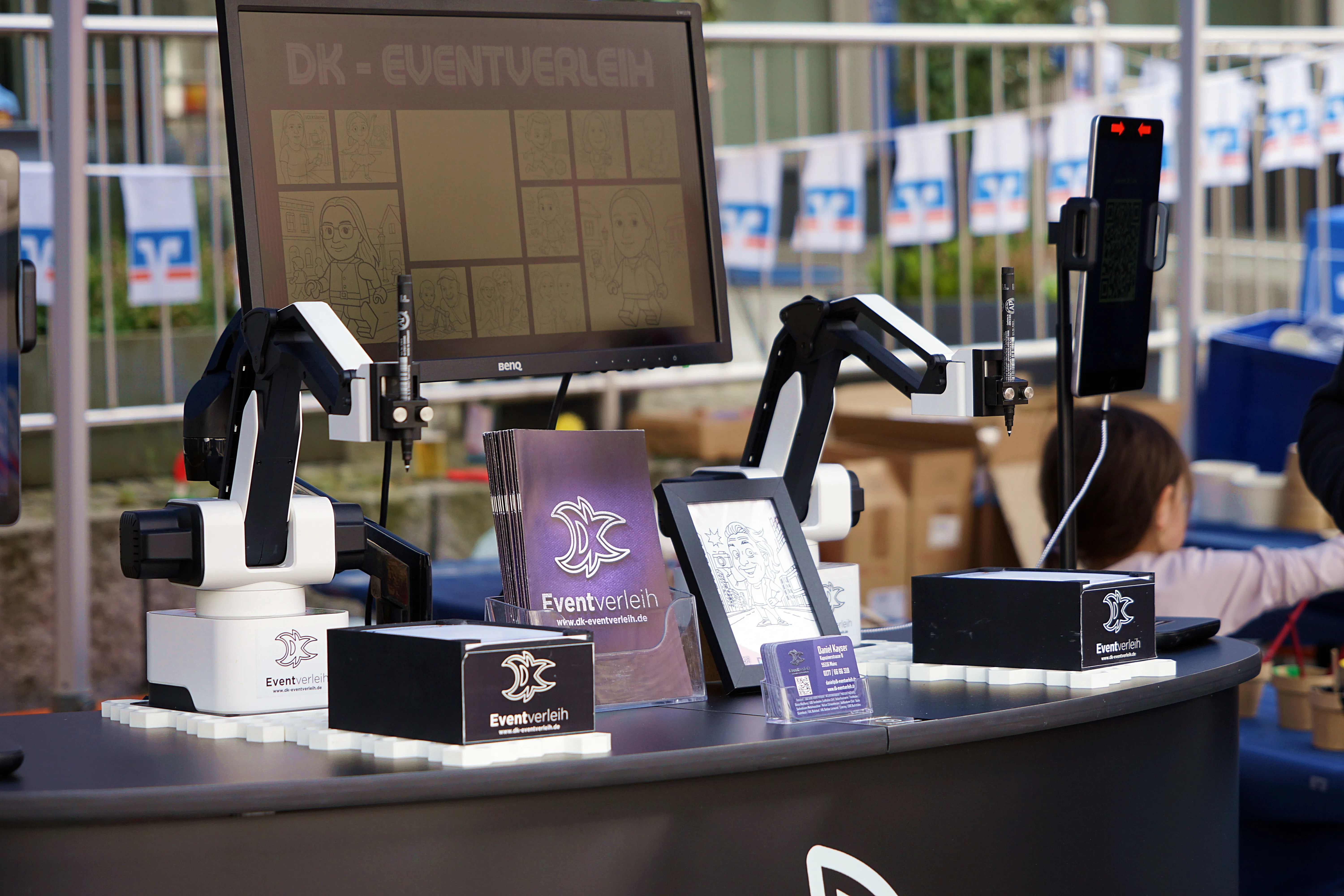 Zwei Roboterarme mit Stiften stehen vor einem Monitor, der Comicfiguren zeigt. Davor liegen Flyer und Visitenkarten des Unternehmens DK-Eventverleih auf einem Tisch.