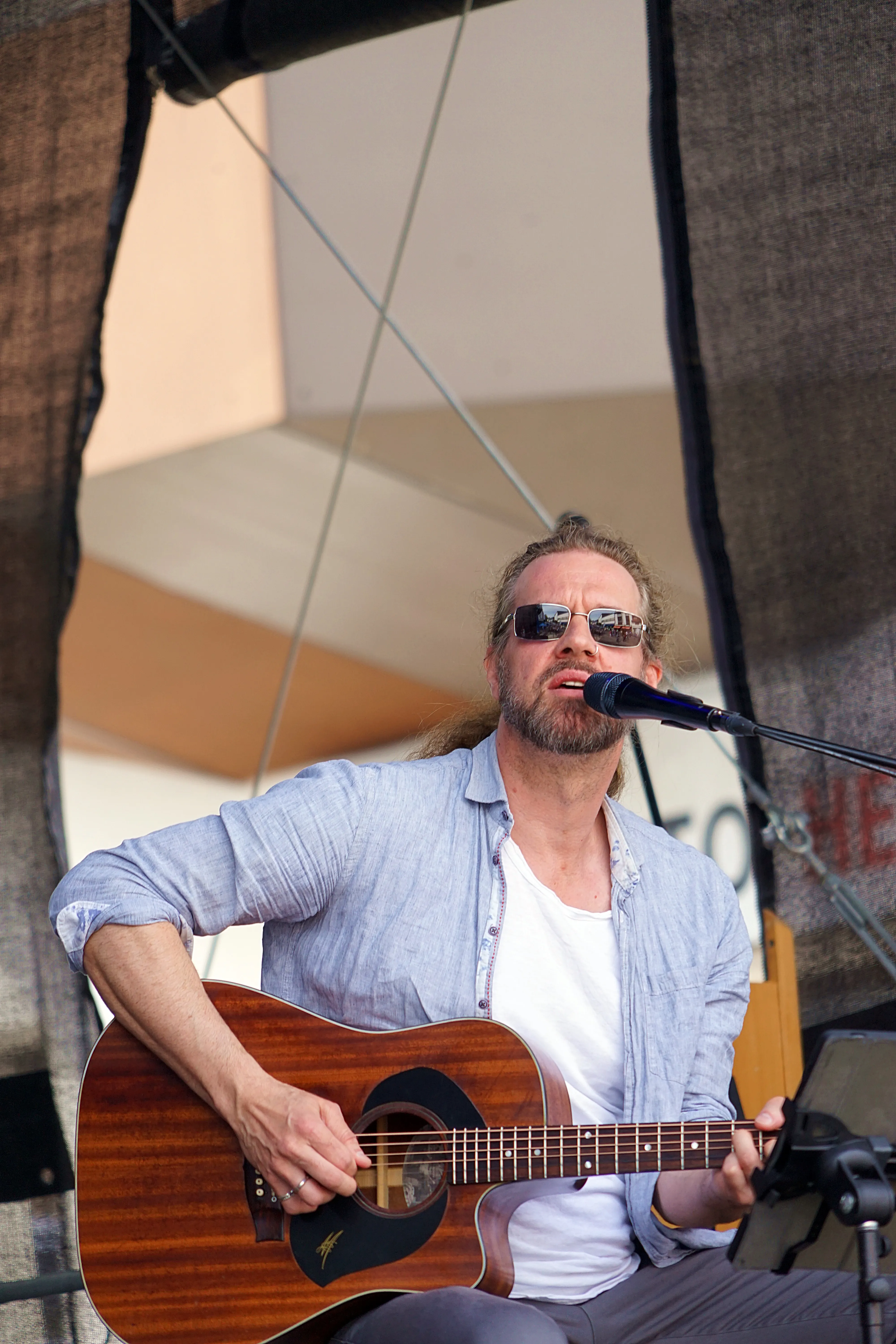 Mann mit Gitarre spielt auf einer Bühne im Freien, trägt Sonnenbrille und helles Hemd.