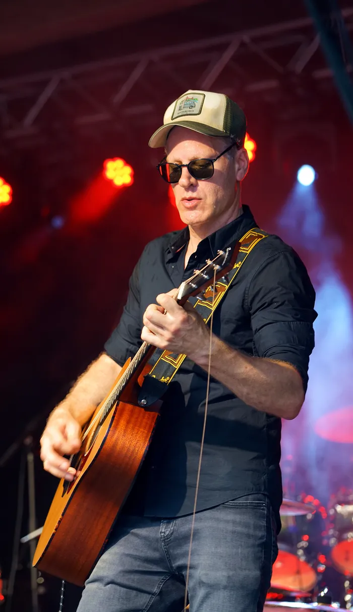 Ein Mann spielt auf einer Akustikgitarre. Er trägt eine Kappe, eine dunkle Bluse und Jeans und hat eine Sonnenbrille auf. Im Hintergrund ist eine Bühne mit roter Beleuchtung zu sehen.