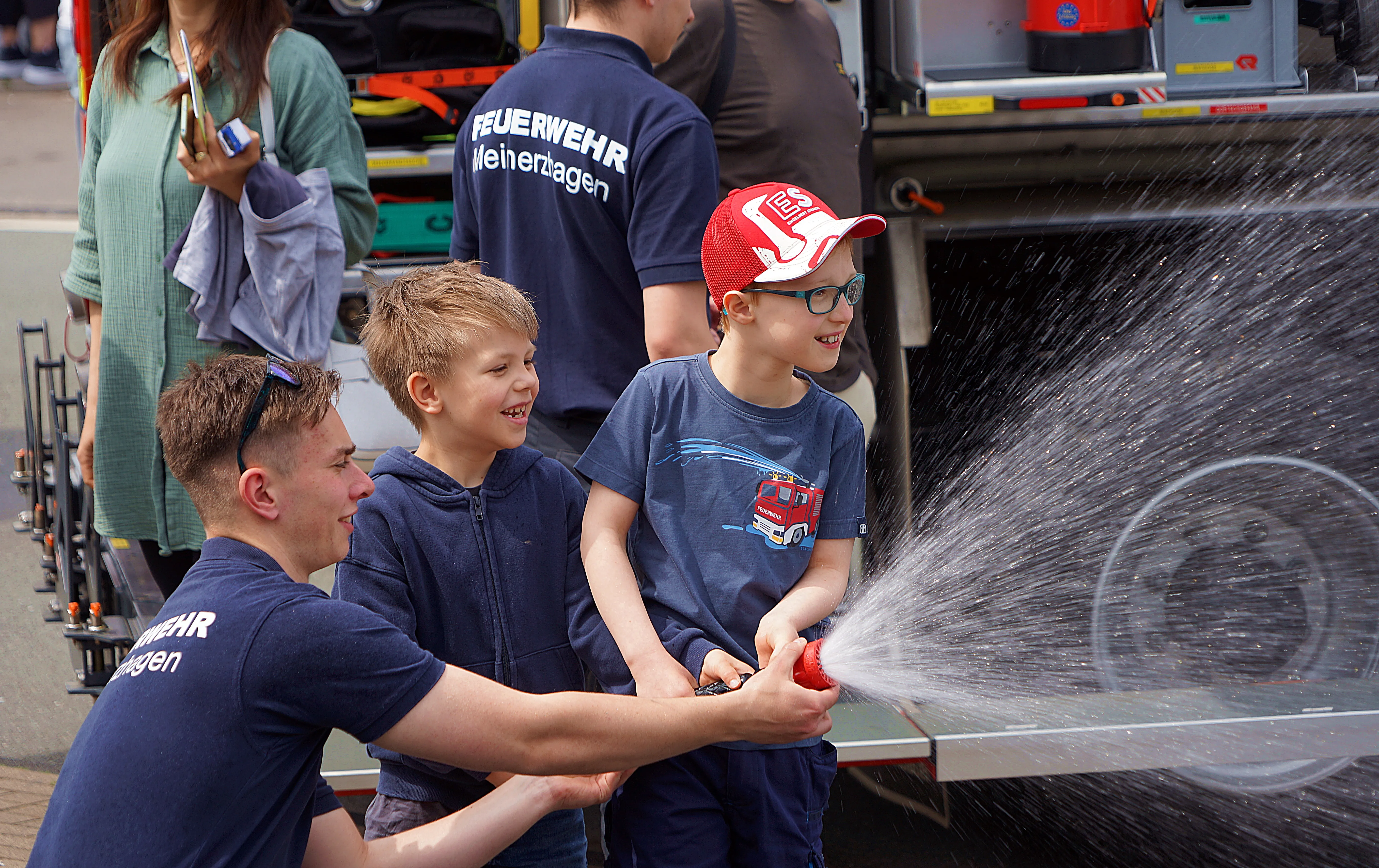 Feuerwehrmann lässt zwei Jungen mit einem Wasserschlauch spielen, während im Hintergrund ein Feuerwehrauto steht. Die Jungen lachen und scheinen Spaß zu haben.