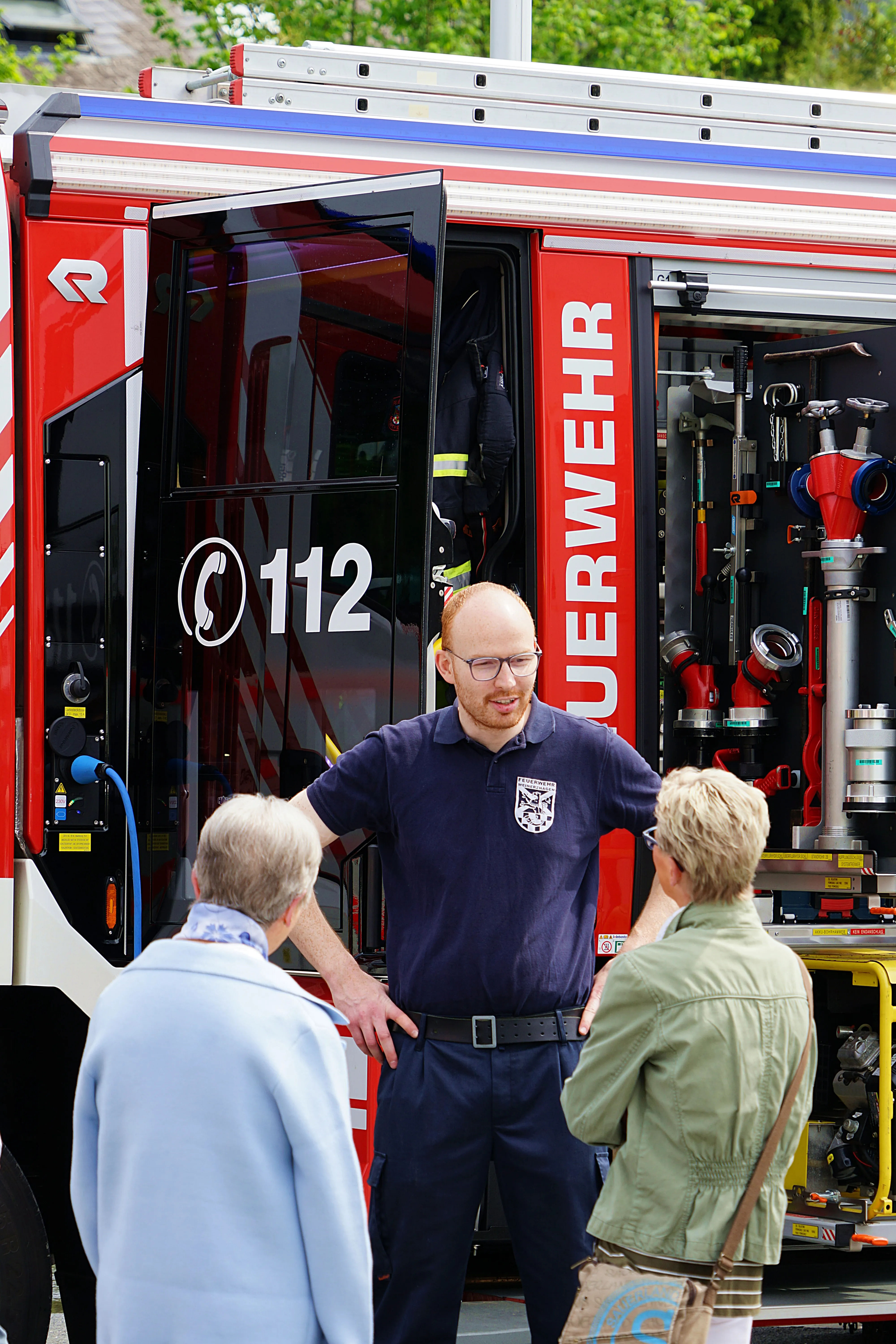 Feuerwehrmann erklärt zwei Frauen und einem Mann etwas vor einem roten Feuerwehrauto mit der Notrufnummer 112.