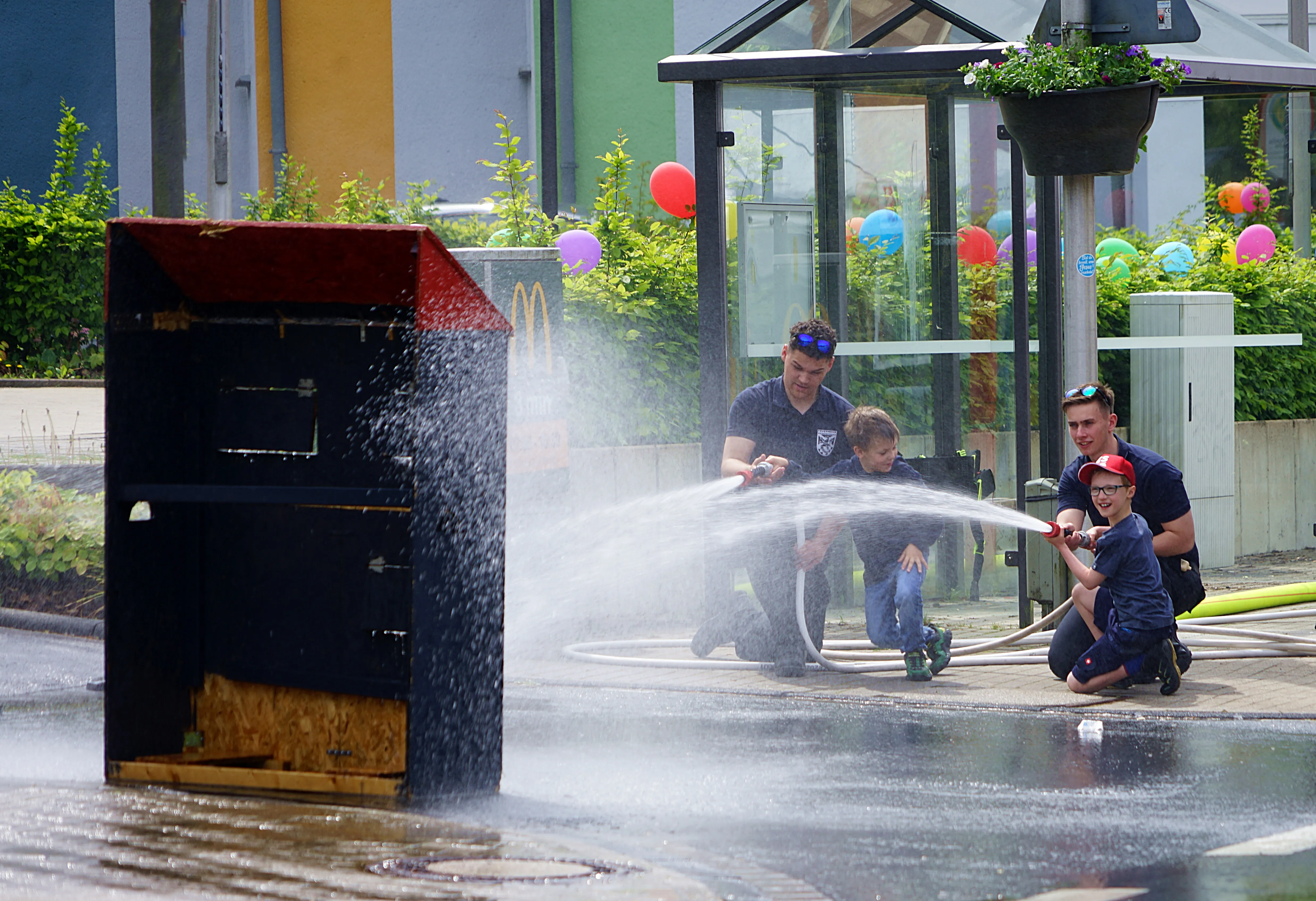 Zwei Erwachsene und zwei Kinder spielen mit Wasserschläuchen vor einer roten Holzkonstruktion und einem Gebäude mit bunten Ballons.