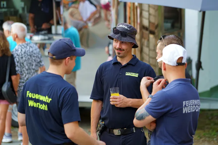 Drei Männer unterhalten sich im Freien. Zwei von ihnen tragen dunkelblaue T-Shirts mit der Aufschrift „Feuerwehr Werohl“, der dritte ein blaues Hemd mit dem Logo der Feuerwehr und eine Eimerhut. Er hält ein Glas mit Orangensaft in der Hand.