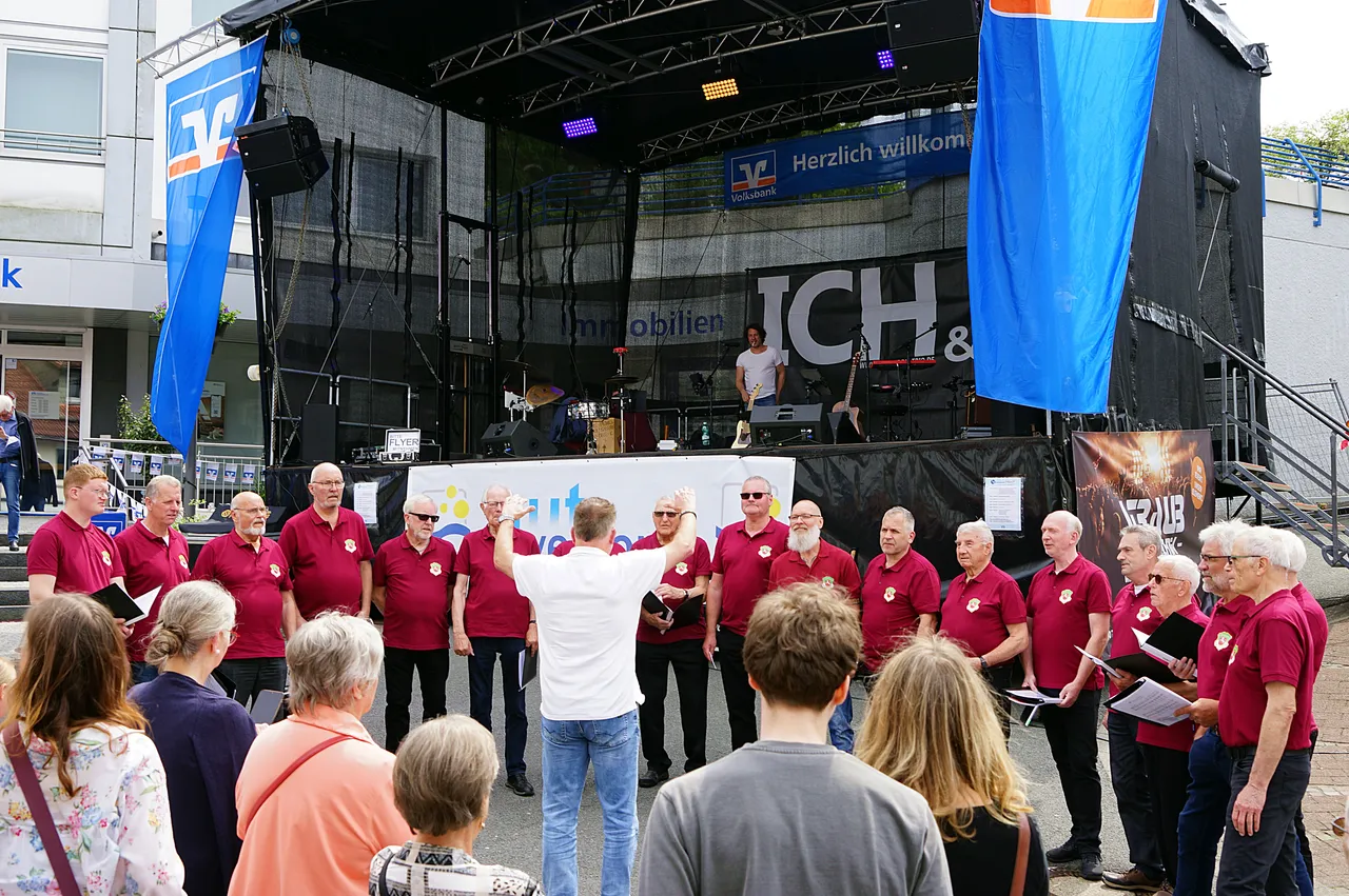 Chor steht auf einem Platz vor einer Bühne mit dem Schriftzug „Immobilien ICH &“ und singt. Zuschauer stehen um den Chor herum und beobachten die Darbietung.