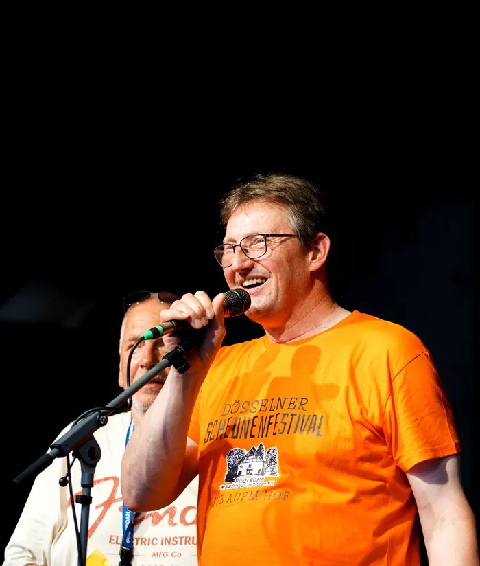 Ein Mann mit rotem T-Shirt und Brille singt in ein Mikrofon auf einer Bühne. Das T-Shirt zeigt den Aufdruck 